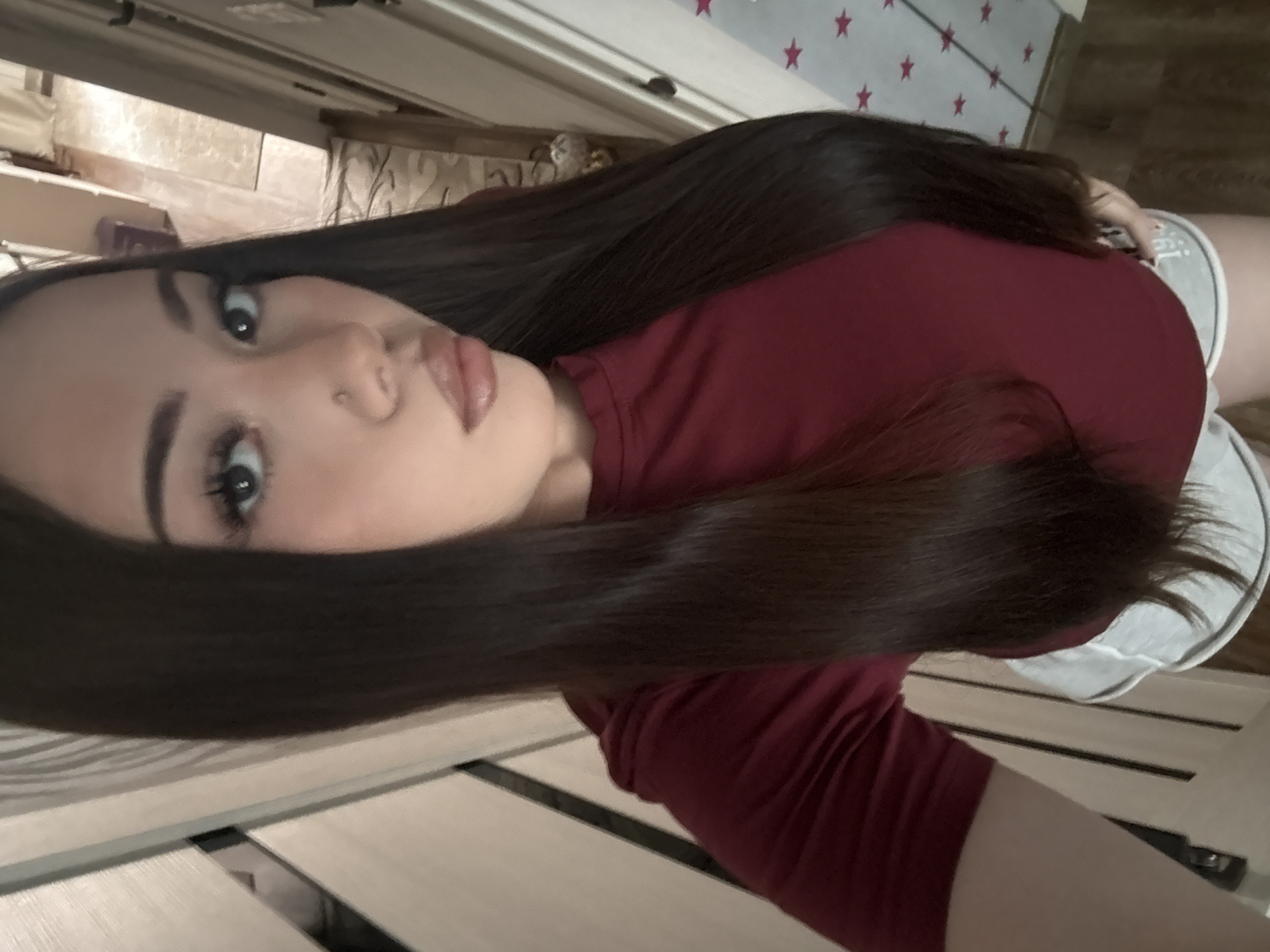 The image is a portrait of a young woman, likely taken indoors, possibly in a bedroom. The photograph is oriented vertically, with the woman's head tilted to her left, giving a rotated perspective.
The subject is a young woman with long, dark, straight hair that cascades over her shoulders and back. She is wearing a dark red, short-sleeved top. Her face features prominent makeup, including defined eyebrows, eyeliner, false eyelashes, and lip gloss or lipstick on her full, slightly pouting lips. A small stud nose piercing is visible on her left nostril. She looks directly towards the camera with a composed expression.
The background reveals a light-colored wall adorned with a repeating pattern of small red stars, visible behind her head and upper body. To the left, another wall with a subtle, possibly patterned, surface can be seen. Below her, a wooden bed frame with visible slats forms part of the setting. On the far right side of the image, a light grey item, possibly part of her lower clothing like shorts or underwear, is partially visible. No discernible text is legible within the image or its provided crops.