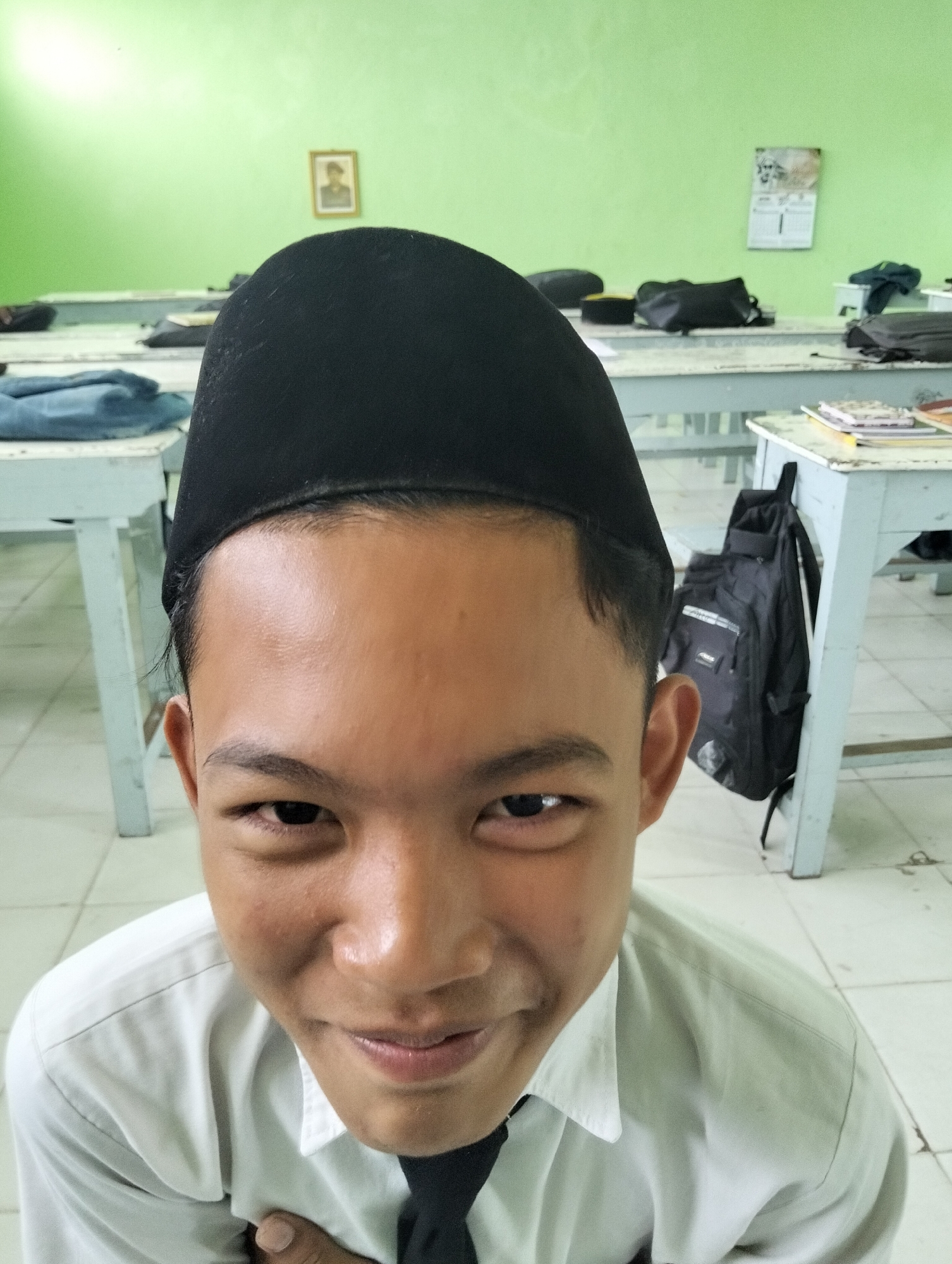 The image captures a close-up, low-angle, selfie-style shot of a young individual wearing a black peci (cap) and a white collared shirt with a dark tie. The subject's face fills the majority of the frame, with a slight smile and direct gaze into the camera. The background suggests a classroom setting with rows of light-colored desks, some with backpacks and books on them. The walls are painted a light green. A framed picture of a person and a calendar are visible on the wall in the background. The floor appears to be tiled. The scene is consistent with a school environment in Kranji, Indonesia.