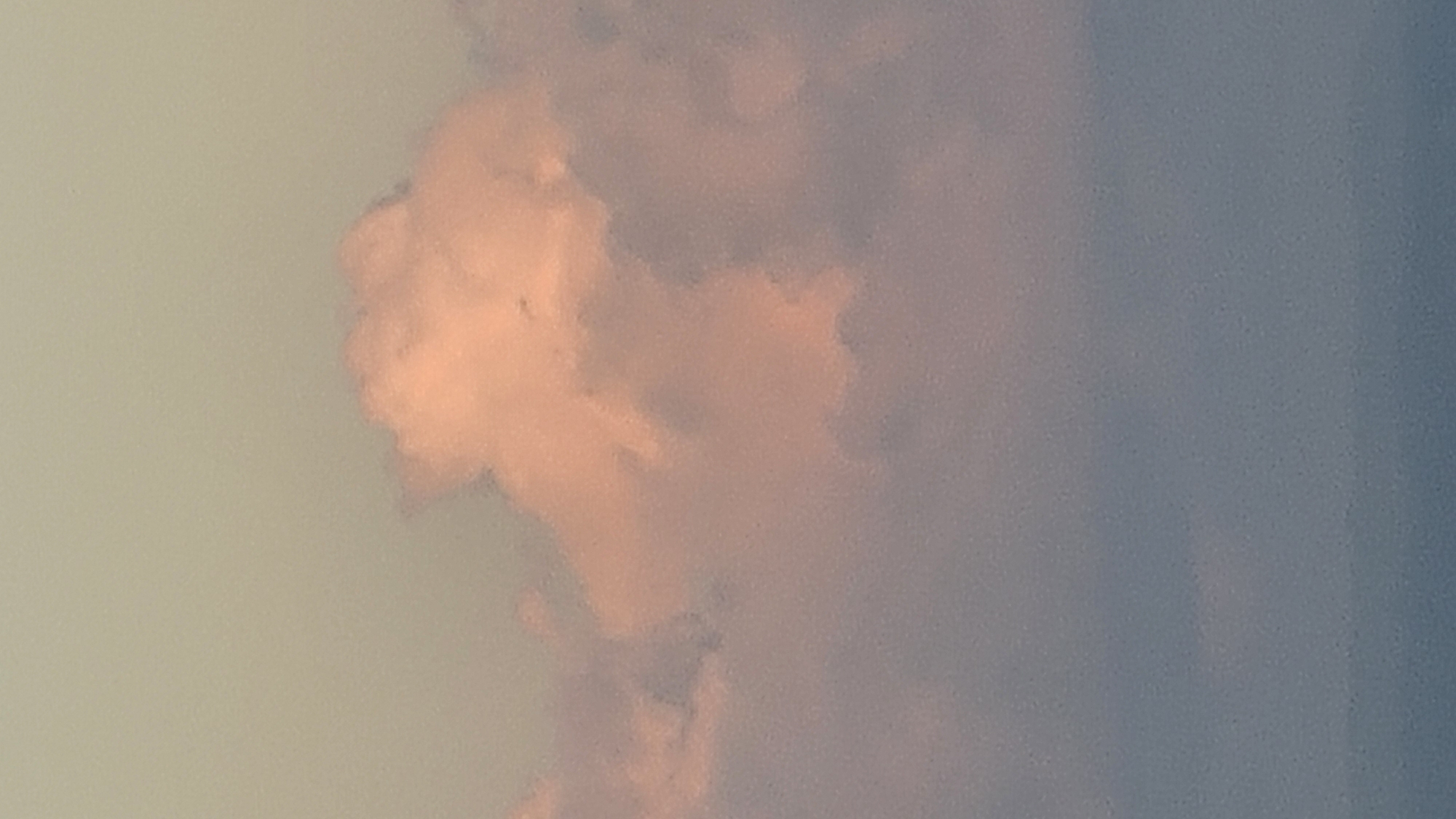 This is a close-up vertical photograph of a pale pink cloud formation against a muted, pale green sky. The cloud is fluffy and textured, with soft edges. Below the pink cloud are layers of darker, purplish-grey clouds, which transition into a pale blue expanse at the bottom of the frame, suggesting the presence of water or a hazy horizon. The image is grainy, characteristic of low-light or older photographic techniques. The scene depicts a natural skyscape, likely during sunrise or sunset, given the color of the clouds. There is no identifiable urban environment or specific location visible within the frame, making it impossible to determine if it is in Denpasar, Indonesia, or any other city.