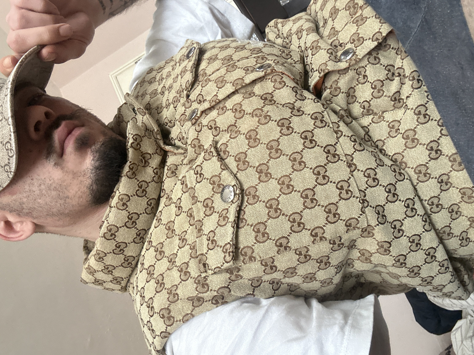 The image presents a close-up, rotated view of a person, appearing to be male, from the chest up. The camera is positioned below the subject, looking upwards, and the image is tilted significantly counter-clockwise, making the person's head appear to be leaning back and looking towards the upper left corner of the frame.
The person has short dark hair and a neatly trimmed beard or stubble covering their jawline and chin. Their lips are slightly parted, and their gaze is directed upwards. Their left hand is raised, with fingers partially obscuring their forehead, holding the brim of a baseball cap. Tattoos are visible on their left forearm and upper arm.
The prominent clothing item is a jacket or thick overshirt, featuring a light beige or tan base color with a repeating brown "GG" monogram pattern, indicative of the Gucci brand. The jacket has several silver-colored snap buttons. Two flapped chest pockets are visible, each secured with one of these snap buttons. A close-up view of one of the buttons reveals engraved text around its circumference: "GUCCI" is visible at the top, and "MADE IN ITALY" is discernible along the bottom curve.
Underneath the monogrammed jacket, a plain white t-shirt or undershirt is visible around the neck and cuffs. On the right side of the main image, a darker fabric, possibly denim or another dark gray garment, is partially visible. The background is simple, consisting primarily of a plain, light-colored wall. The overall scene captures a casual moment, emphasizing the person's distinctive branded attire.