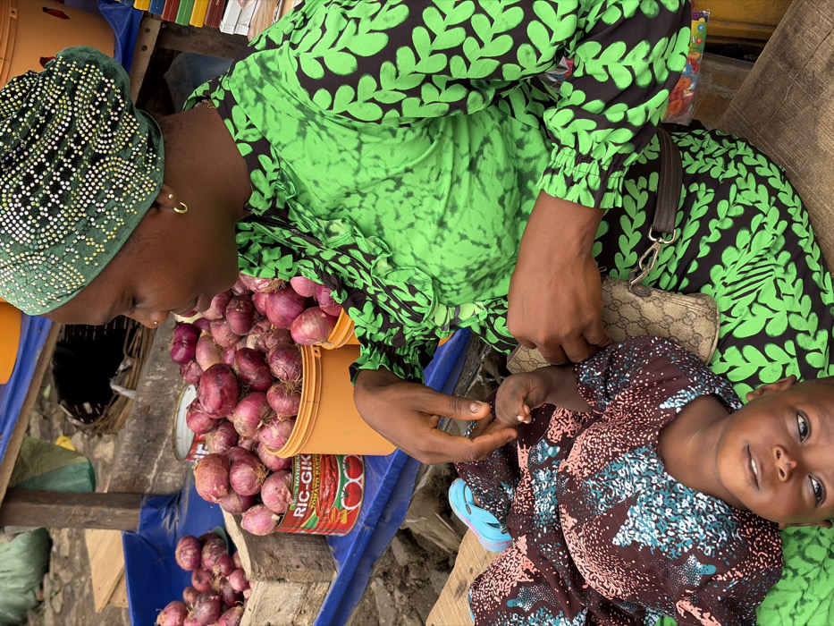 The image depicts an adult female and a child at an outdoor market stall in Jakande, Nigeria. The adult, with dark skin, is seated and wears a long-sleeved green dress featuring black leaf patterns and ruffles on the bodice. She also wears a green headwrap adorned with metallic studs in a geometric pattern, small gold hoop earrings, and a nose stud. Her right hand holds the left hand of a child lying on its back on a surface adjacent to her. The child, dressed in a brown, pink, and light blue patterned outfit, looks upward towards the adult; a single light blue sandal is visible on its left foot. A small, patterned purse, appearing to be a designer brand, is attached to a brown belt-like strap and rests on the adult's lap. Behind the adult, a market stall displays various quantities of red onions. These onions are contained within an orange plastic bucket, a wooden crate, and a red metal can labeled "RIC-Gik 100% Natural Tomato Paste," all situated on a blue surface. The ground visible in the background is indistinct and appears to be an uneven dirt or concrete surface.