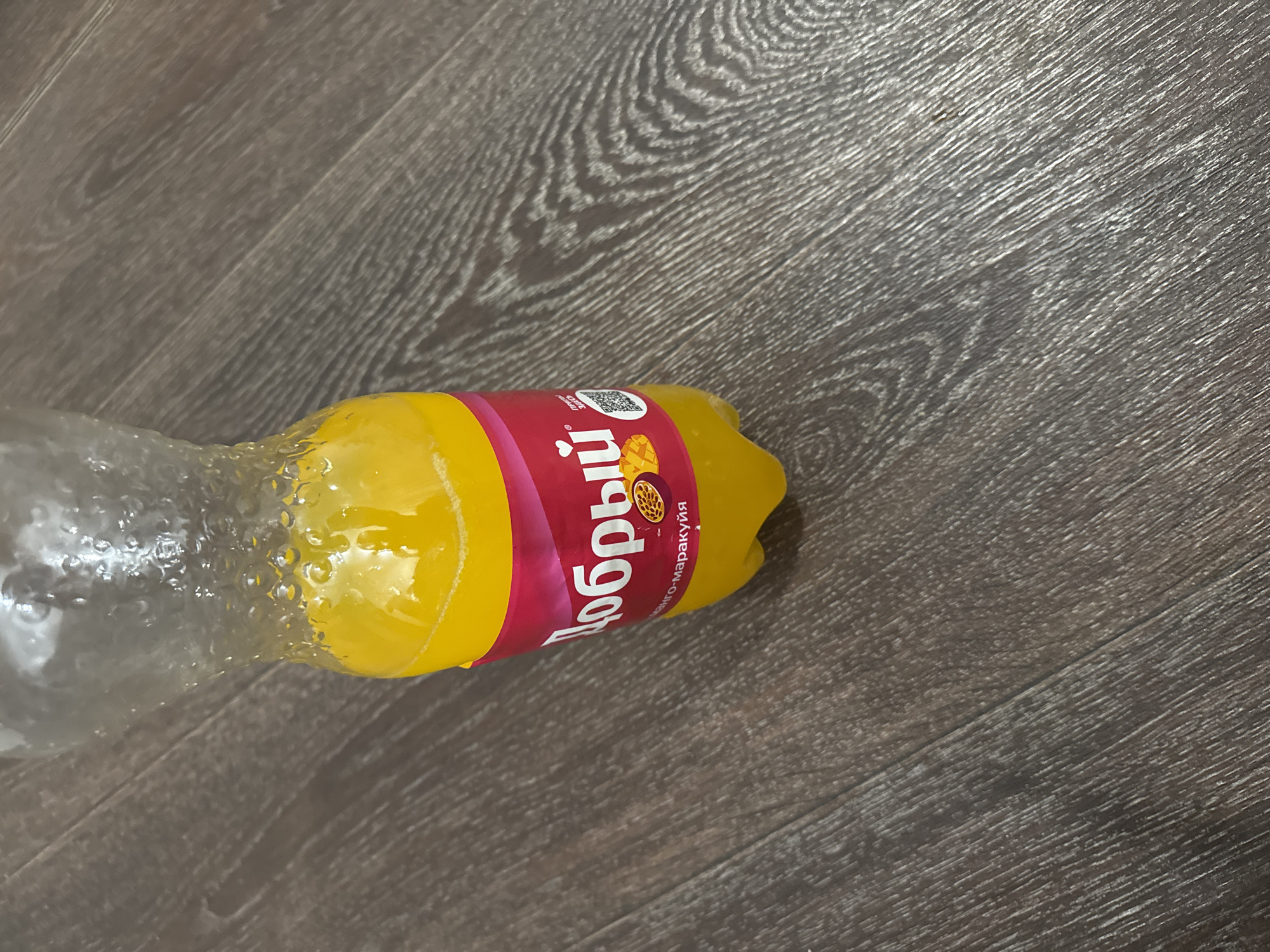 A plastic bottle with yellow liquid and a label is resting on a wooden floor. The label is red and white with Cyrillic text and an image of passion fruit and mango. The bottle is partially filled with the liquid, and condensation is visible on the upper portion of the bottle. The wooden floor has a dark brown color and a visible wood grain pattern. There are no people or other objects in the immediate vicinity. The scene appears to be indoors, possibly in a home or commercial establishment. The lighting is moderate, and the image is in focus.