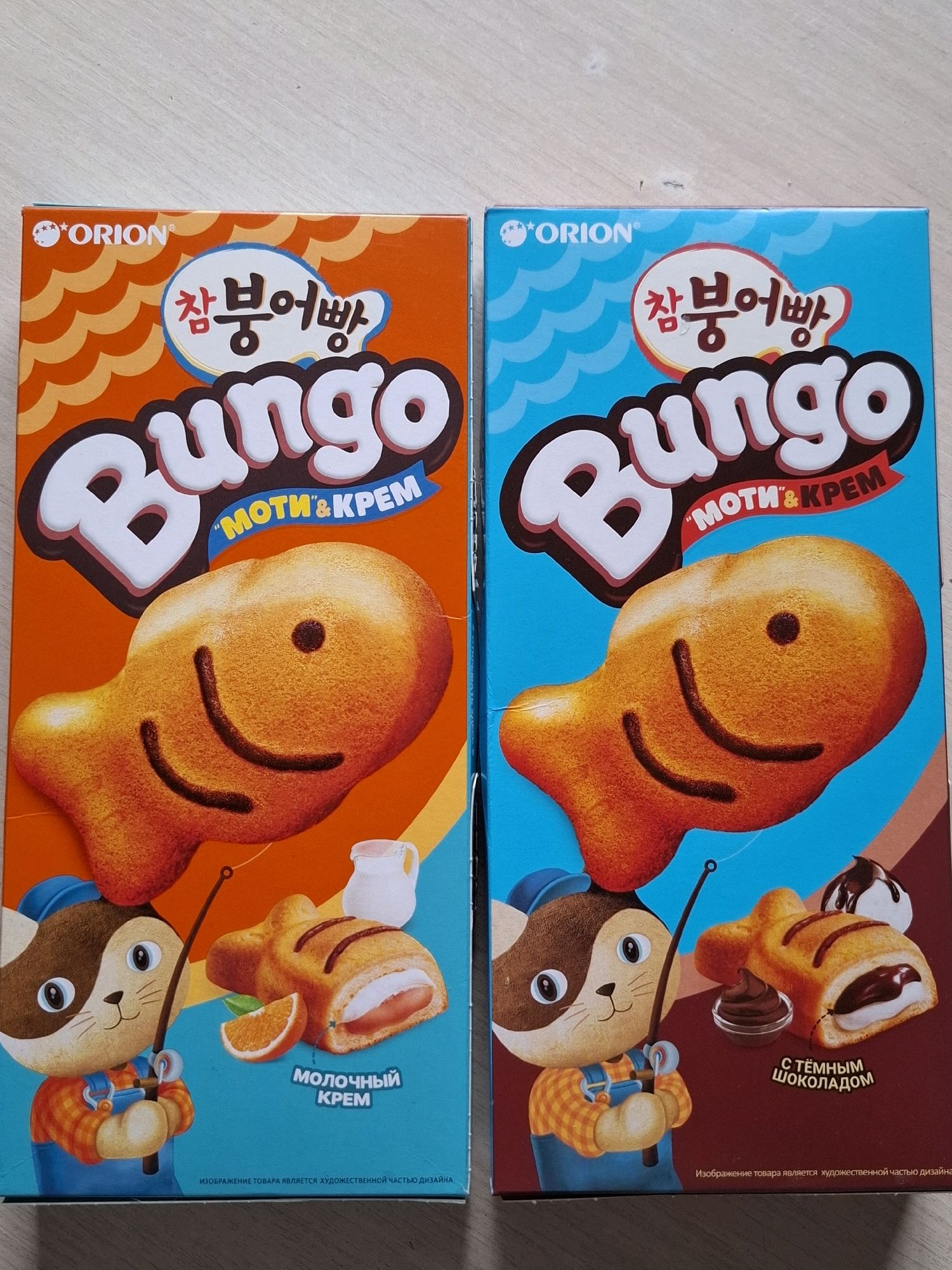 The image displays two boxes of Orion Bungeo-ppang snacks. The box on the left has an orange background and features a smiling fish-shaped snack with cream filling and an orange slice. A cartoon cat with a fishing rod is also depicted. The box on the right has a blue and brown background and shows a similar fish-shaped snack with chocolate filling and a dollop of chocolate sauce. The cat character is also present on this box. The text on the boxes is in Korean and Russian, indicating "Moti & Krem" (cream) and "with dark chocolate" in Russian. The overall presentation suggests a product marketed towards children, with bright colors and playful imagery. The location of this image in Tyumen, Russia cannot be determined from the visual information provided.