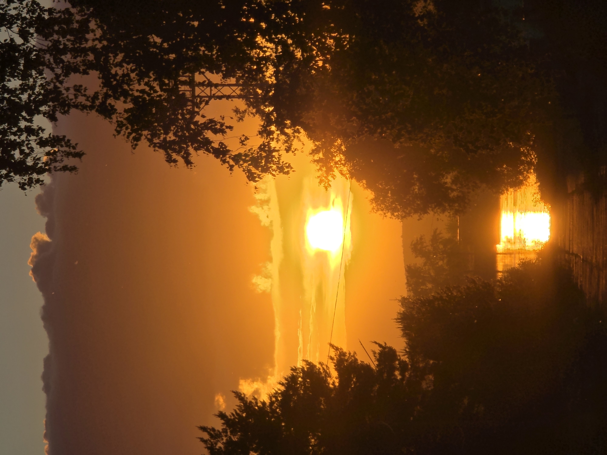 The image depicts a dramatic sunrise or sunset in Chaikovsky, Russia, characterized by an overwhelming golden and orange light. The sun, a brilliant white sphere, is centrally located towards the left side of the frame, emitting an intense glow that bathes the entire sky in fiery hues.
Silhouetted dark green foliage from trees and bushes frames the upper and right portions of the scene, with individual leaves catching the backlighting. A large, dark cloud formation stretches across the bottom edge of the sky, contrasting with the radiant light. A power line or cable is visible, running vertically downwards through the intensely lit area.
In the distance on the right, a building or large structure is heavily backlit, appearing as a bright, reflective rectangular shape, possibly with windows, catching the sun's powerful rays. The atmosphere appears hazy, enhancing the vibrant colors and diffusion of light. No people or visible text are present. The scene evokes a strong sense of natural beauty and awe due to the spectacular lighting conditions.