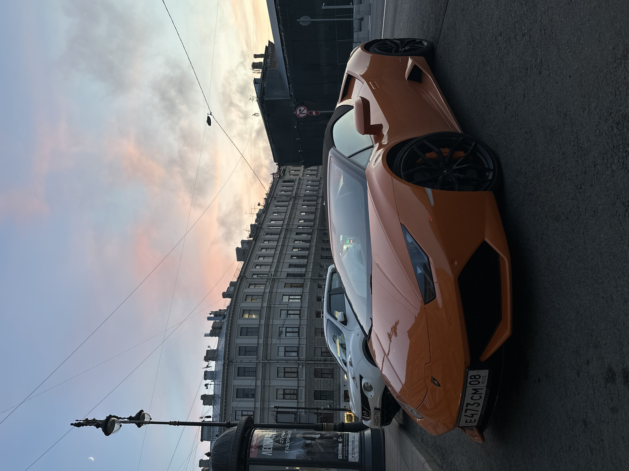 A tilted, ground-level shot shows two parked cars on a street in St. Petersburg, Russia, with a twilight sky in the background. The prominent vehicle is an orange Lamborghini, partially obstructing a white car behind it. A street lamp and an advertisement kiosk are visible on the left, close to the edge of the frame. The backdrop is a row of buildings, typical of the city's architecture, under a sky with pink and blue hues from the setting sun. The advertisement kiosk exhibits Russian text, including "ЛЕОНИД" and "ГАЗПРОМА". The license plate of the Lamborghini reads "E473 CM 08 RUS". A “Royal Thai” advertisement appears on the kiosk as well. A crescent moon is visible in the sky. Overhead wires traverse the sky, adding to the urban environment.