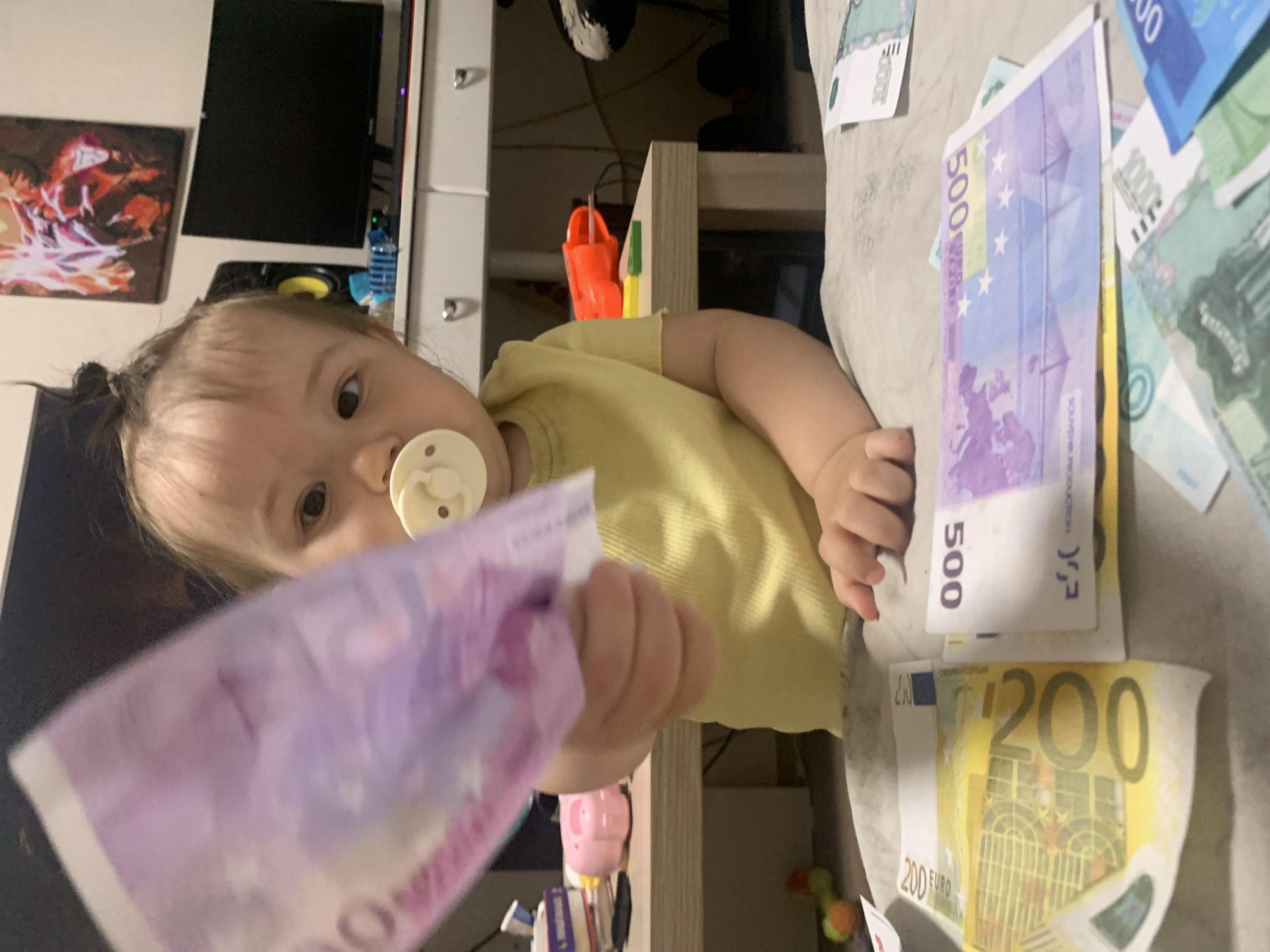 A baby wearing a yellow shirt and a pacifier is holding a 500 euro banknote. In front of the baby are several other banknotes, including a 200 euro banknote and a 1000 euro banknote. The background shows a desk with computers and other objects. The scene is indoors, likely in a residential setting. The city of Chaikovsky, Russia is not depicted in this image.