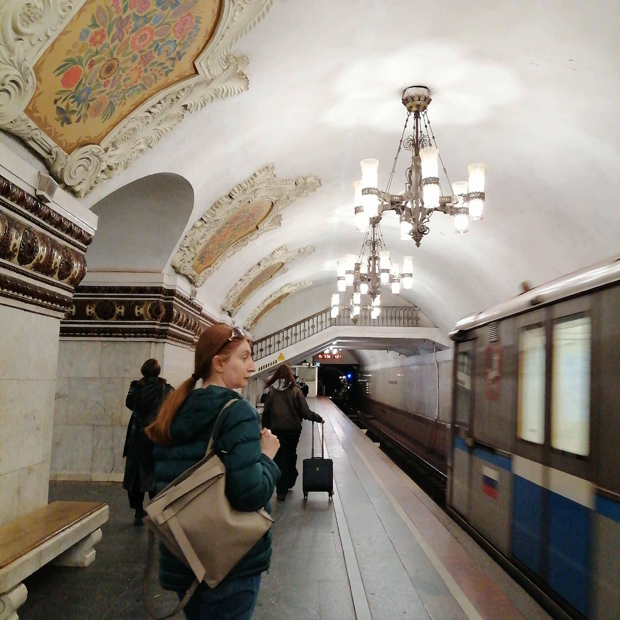 The image captures a scene within the Moscow Metro, Russia. A subway train, marked with a Russian flag emblem, is moving alongside the platform. A woman, identifiable by her red hair pulled into a ponytail and wearing a green jacket and jeans, stands on the platform with a shoulder bag. Another individual is walking in the same direction, pulling a wheeled suitcase. A third person is slightly behind them. The subway station exhibits ornate architectural details including chandeliers, decorated arches, marble columns, and a ceiling with floral patterns.