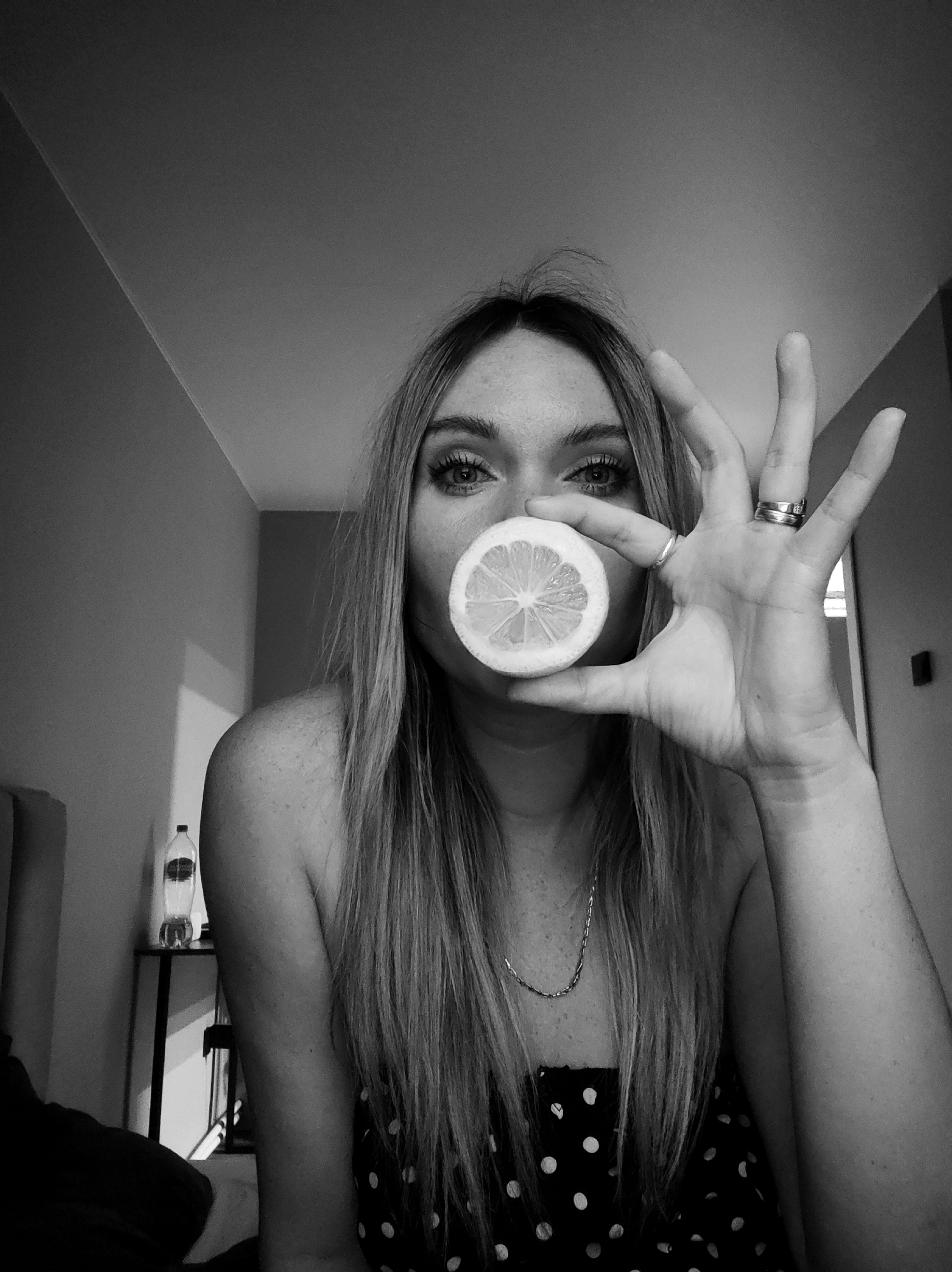 A black and white photograph shows a woman indoors holding a lemon slice to her mouth with her hand. The woman has long hair, and is wearing a necklace, rings, and a polka dot top. A bottle of water sits on a table in the background. The location is unknown, but is not believed to be in Votkinsk, Russia.