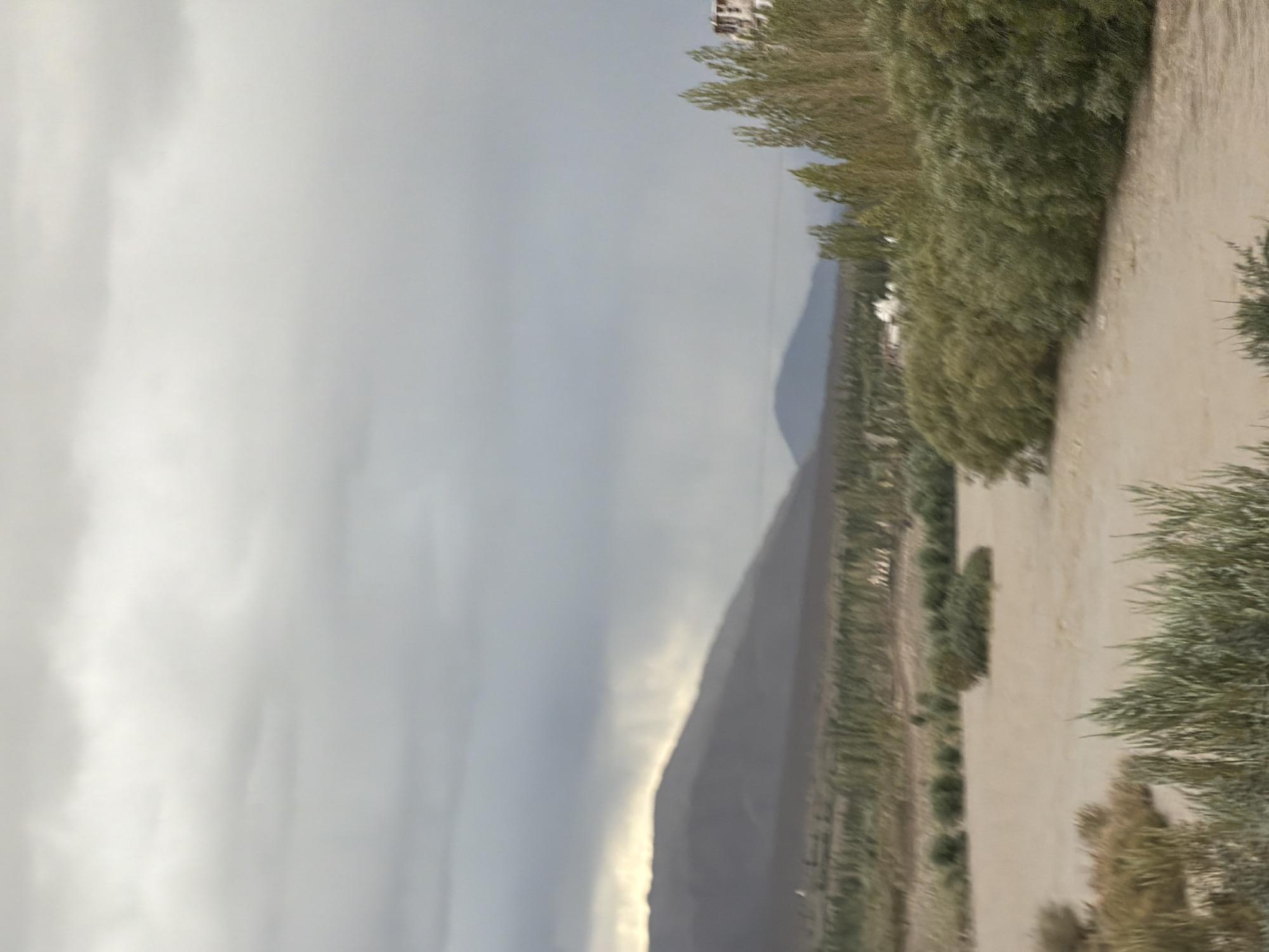 An overcast sky looms above a wide, muddy river flowing through a valley. The riverbanks are lined with lush green vegetation, including trees and bushes, some of which appear dense and overgrown. In the background, rolling hills or mountains are visible, their slopes partially obscured by the mist or low-hanging clouds. On the right side of the image, a cluster of white buildings with a red roof is nestled among the trees, suggesting a settlement or village. The overall scene depicts a natural landscape that is somewhat undeveloped, with a hint of human habitation. The location is identified as Tanglakpurna, India.