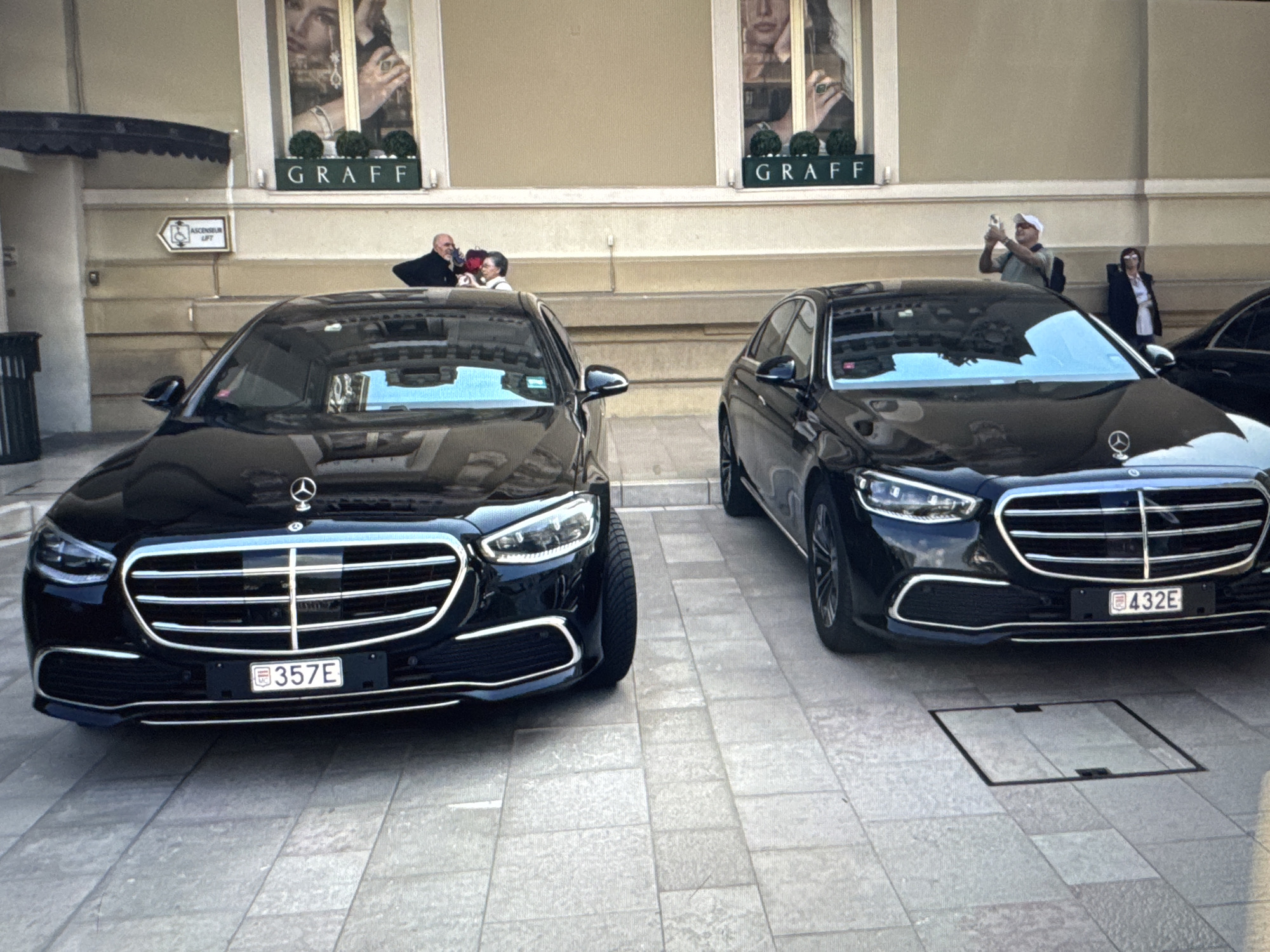 Two black Mercedes-Benz S-Class sedans are parked side-by-side on a tiled pedestrian walkway. The car on the left has license plate "357E" and the car on the right has license plate "432E". In the background, a building with large display windows for "GRAFF" jewelry is visible. Several people are in front of the building, including one individual taking a photograph with a cell phone. There is a sign pointing to an elevator. The scene appears to be in an urban environment, likely a commercial or luxury district.