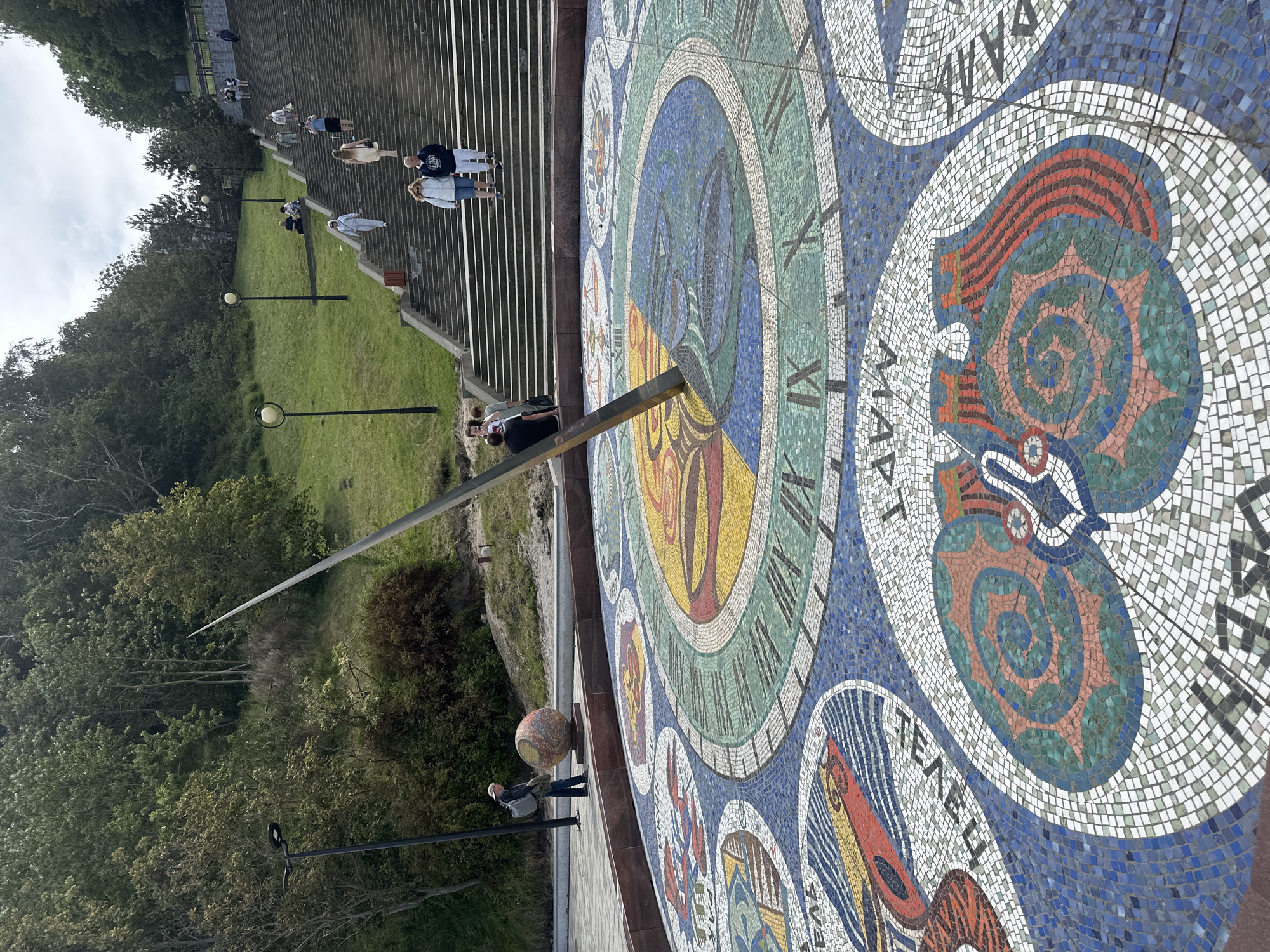 In Svetlogorsk, Russia, a large, ornate sundial mosaic dominates the foreground of this outdoor scene. The sundial is circular, composed of intricate mosaics featuring various patterns and colors, including stylized representations of celestial bodies and zodiac signs. A metal gnomon, casting a shadow, is centrally positioned on the sundial.
Behind the sundial, a wide, grand staircase ascends a grassy hill. Several people are visible on the stairs, some walking up or down, while others are seated on the steps. The hill is covered in lush green grass and trees, creating a natural backdrop.
To the left of the sundial, a man stands near a spherical, mosaic-covered sculpture. A lamppost is also present in this area. The overall atmosphere suggests a public park or recreational area, with people enjoying the outdoors and interacting with the artwork and the natural environment. The sky is overcast.