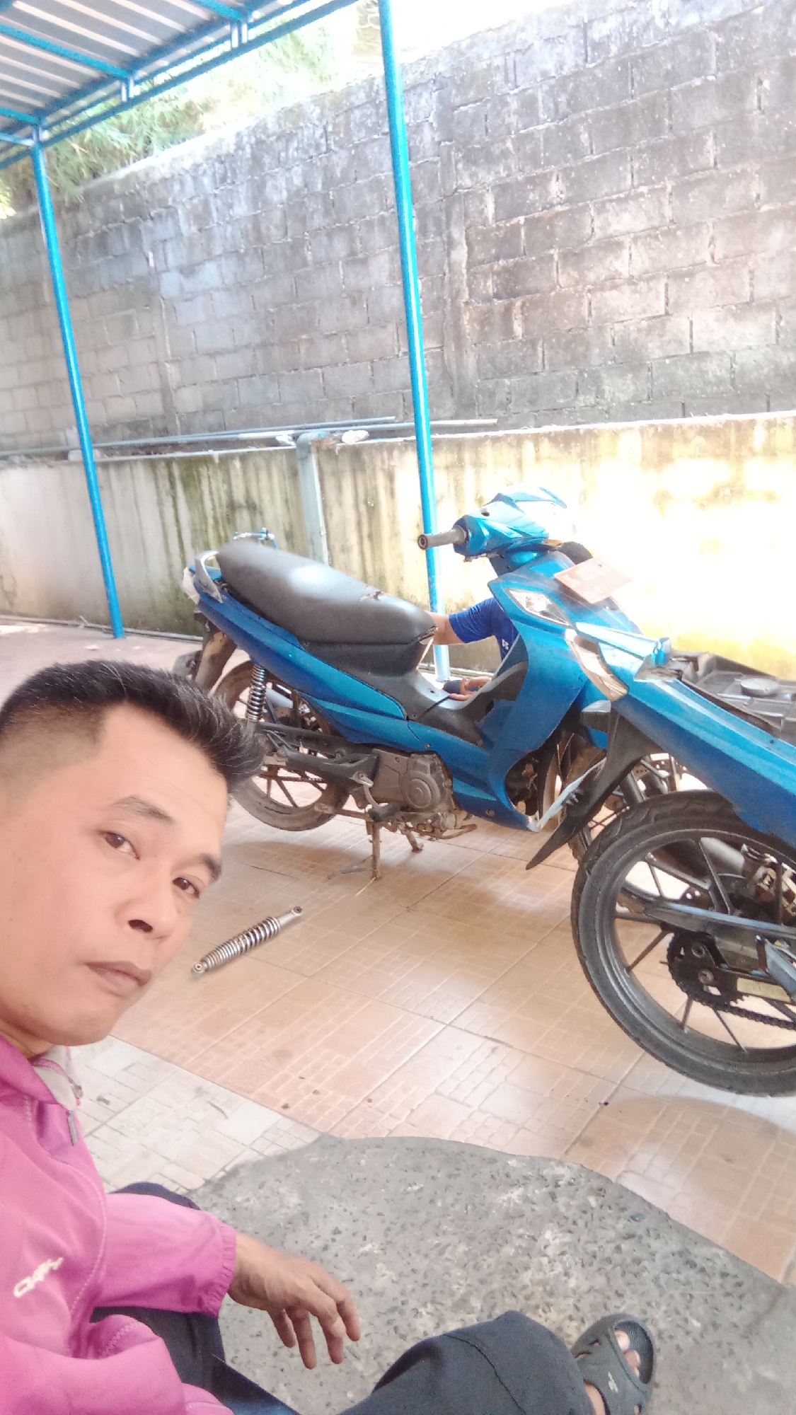 A man in a pink jacket and dark pants sits on the ground next to a blue motorcycle. The motorcycle is parked on a tiled floor under a covered structure. The structure's roof is corrugated metal, supported by blue poles. Behind the motorcycle is a brick wall. A spare part, possibly a shock absorber, lies on the ground near the man's left hand. The man is looking towards the camera. The scene appears to be an outdoor workshop or garage in Jawa, Indonesia, possibly during daylight hours.