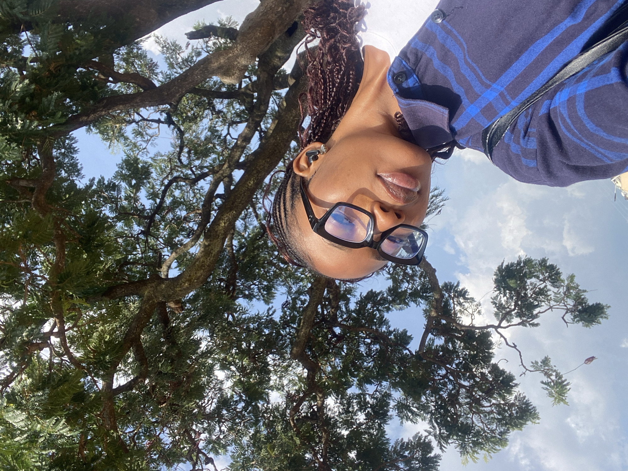 A low-angle, outdoor shot captures a young Black woman with braided hair and black-rimmed glasses. She is wearing a blue and black plaid shirt. She is looking up and to the side, her lips slightly parted, and she has an earbud in her left ear. Behind her, a large tree with green leaves and a cloudy blue sky are visible. The scene appears to be in Jos, Nigeria, based on the overall context of the image.