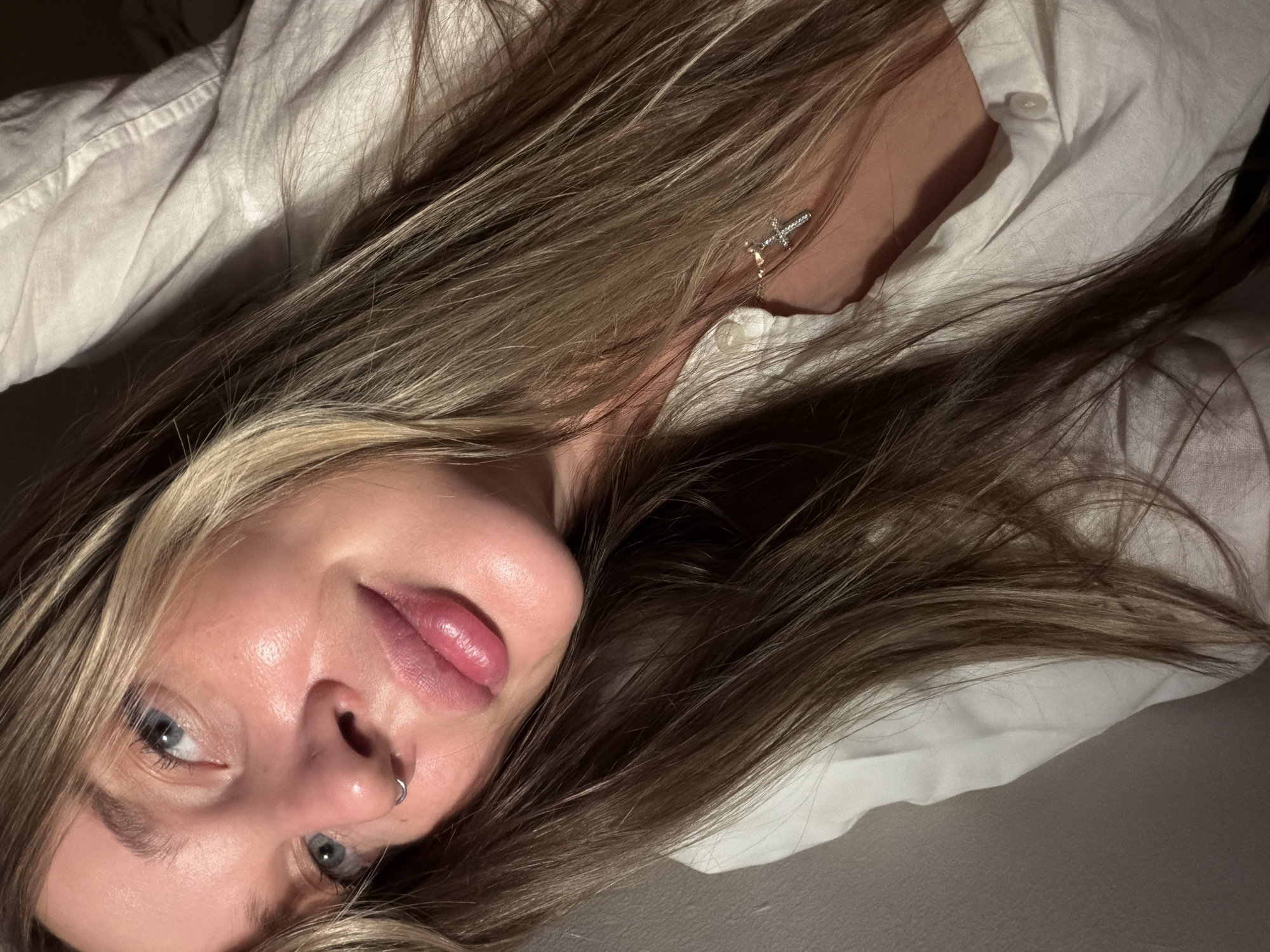 A selfie taken from a low angle features a young woman with blonde hair, wearing a white button-down shirt. She has blue eyes and a nose piercing. Her face is well-lit and angled towards the camera. A silver cross earring is visible. The background shows a white surface and the fabric of her shirt. The image appears to be taken indoors. There is no information to suggest that the image was taken in Moscow, Russia.