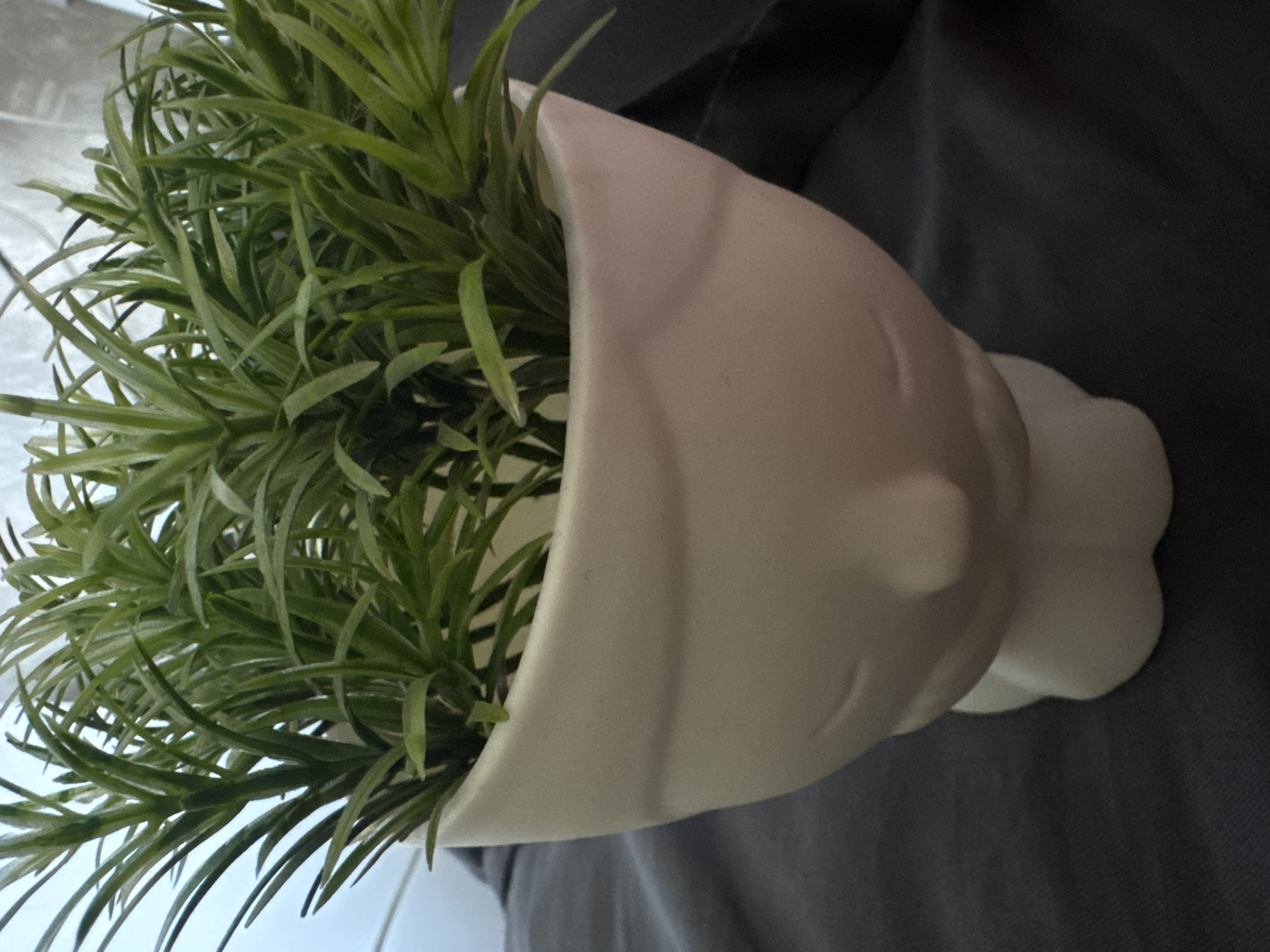 The image shows a close-up of a white ceramic planter shaped like a face, holding a bunch of green artificial plants. The planter has closed eyes and a small nose, with its base resembling the chin and neck. It rests on a dark grey fabric surface, likely a tablecloth or blanket. The background is blurred, with hints of a light-colored wall and possibly a window. The overall lighting suggests an indoor setting. This scene is not located in Sofia, Bulgaria, but rather appears to be a still life arrangement in a private residence or studio.