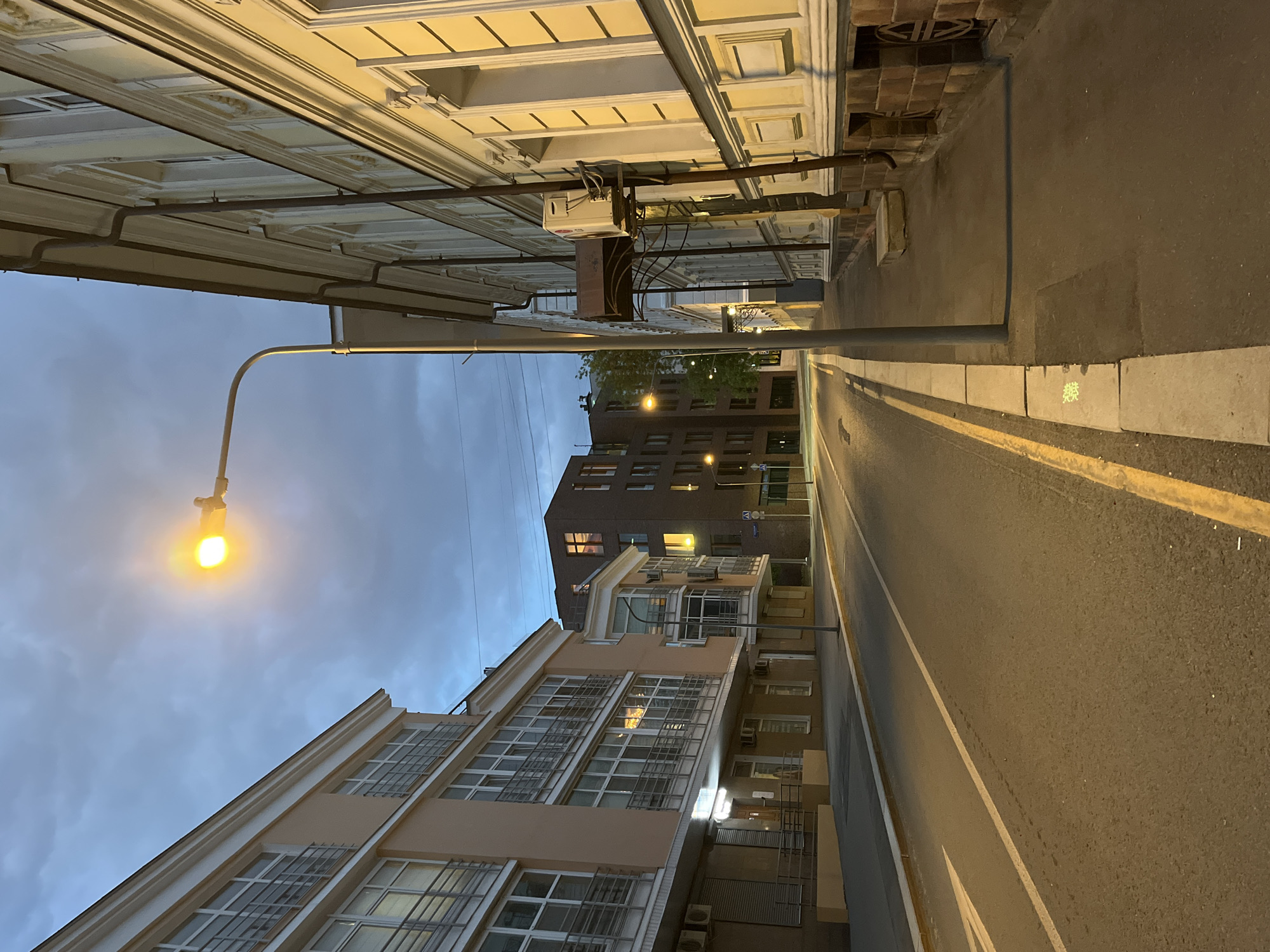 The image depicts a city street at dusk. Buildings flank both sides of the road. The building on the left is a lighter color, possibly beige or cream, with numerous windows, some of which have metal grilles. The building on the right is darker, possibly brown or brick, and also has several windows. A street lamp on the left illuminates the scene with a warm yellow light. The road has yellow lines marking the lanes and curbing. A manhole cover is visible on the right. The sky is a mix of light blue and gray, suggesting the time is either early morning or late evening. The general location appears to be an urban setting.