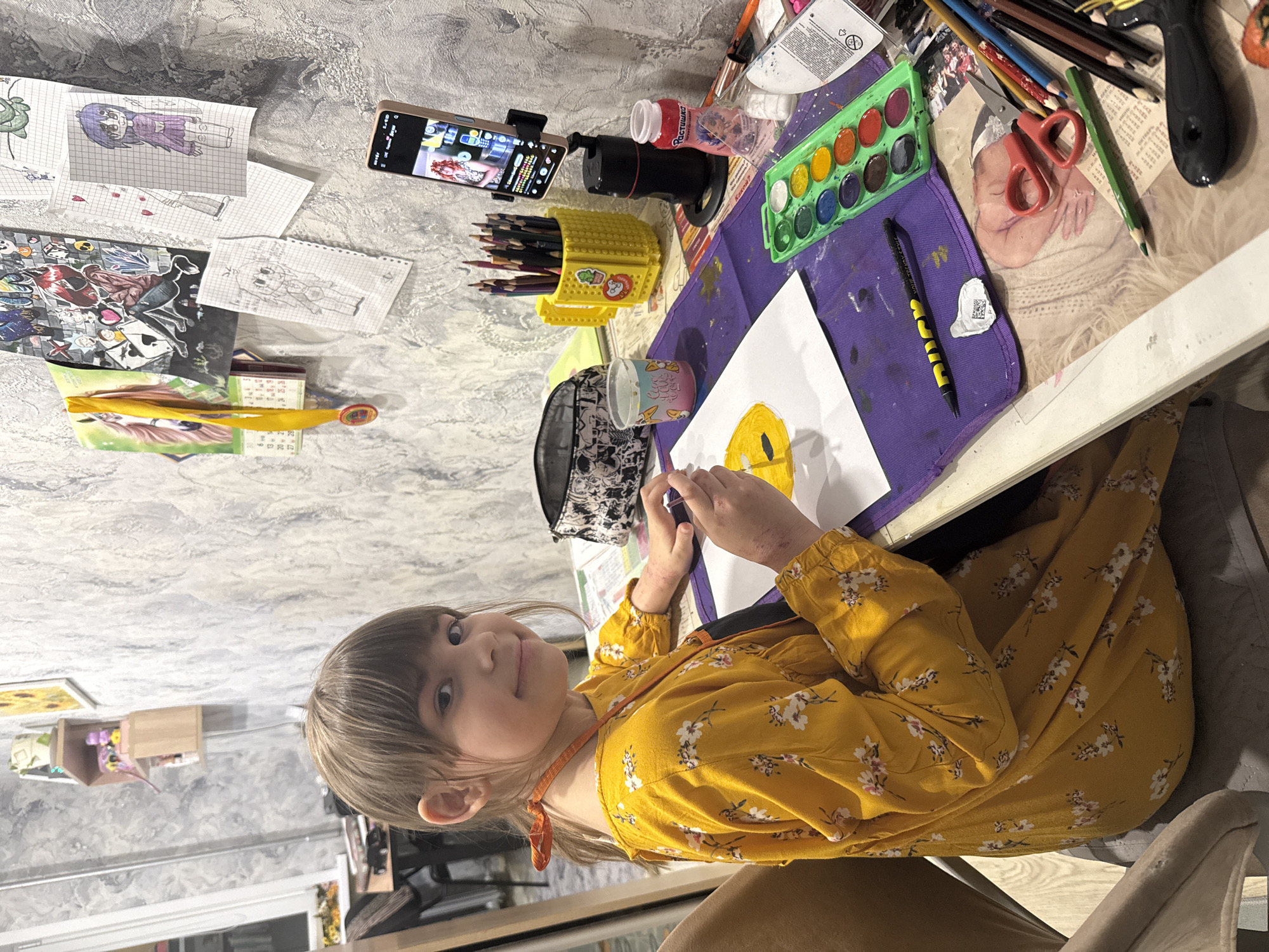 A young girl, likely of early elementary age, is pictured seated at a table, engaged in an art activity. She has light brown hair with bangs and is wearing a long-sleeved yellow dress with a white floral pattern and an orange cord around her neck. She looks towards the camera with a slight smile. Her left hand holds a paintbrush, working on a white sheet of paper with a partially painted yellow circular shape. Both her hands show some red markings.
The table is filled with various art supplies. To the girl's left, a smartphone, held in a black stand, displays a video or image of a woman with red curly hair, suggesting she might be following an online tutorial or watching entertainment. Adjacent to the phone are a black device and a yellow Lego-style container holding numerous colored pencils. A green-cased watercolor set, additional pencils, a black pen with "DUCH" visible on its yellow barrel, and a clear cup containing water for painting are also on the table. A plastic bottle labeled "Растушка" (Rastushka) in Cyrillic and a white tube of paint with Cyrillic text are present. A purple mat, stained with paint, is beneath her painting area.
Other items on the table and wall include several hand-drawn anime/manga style characters on grid paper (one with purple hair, another a turtle, and one with two characters holding hands), a larger print with various characters, a small calendar tied with a yellow ribbon, red-handled scissors, and a framed photograph of a sleeping baby.
The background shows a wall with textured grey and white wallpaper. Further back, shelves and a framed sunflower painting are visible. The scene depicts a child's creative art session at home.