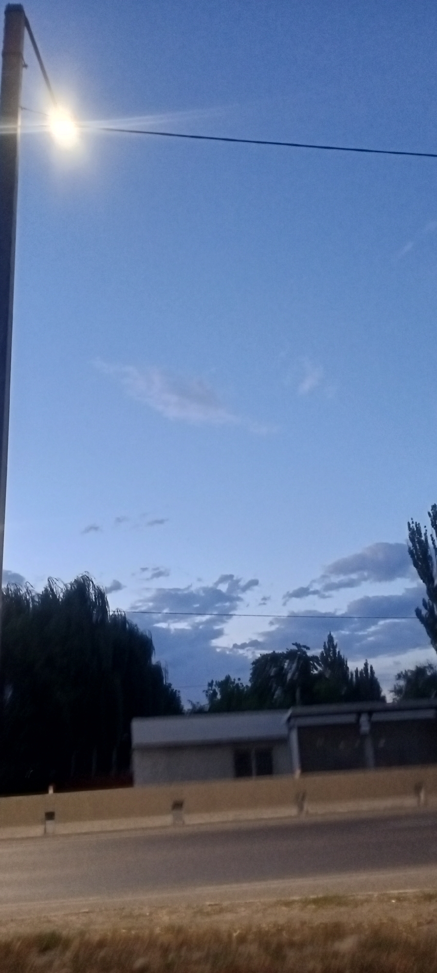 A street lamp illuminates a vast, clear blue sky with scattered white clouds. In the distance, silhouetted trees and a low-lying building are visible, bordered by a highway with a concrete barrier. The image appears to have been taken at dusk in Romanovka, Kyrgyzstan.