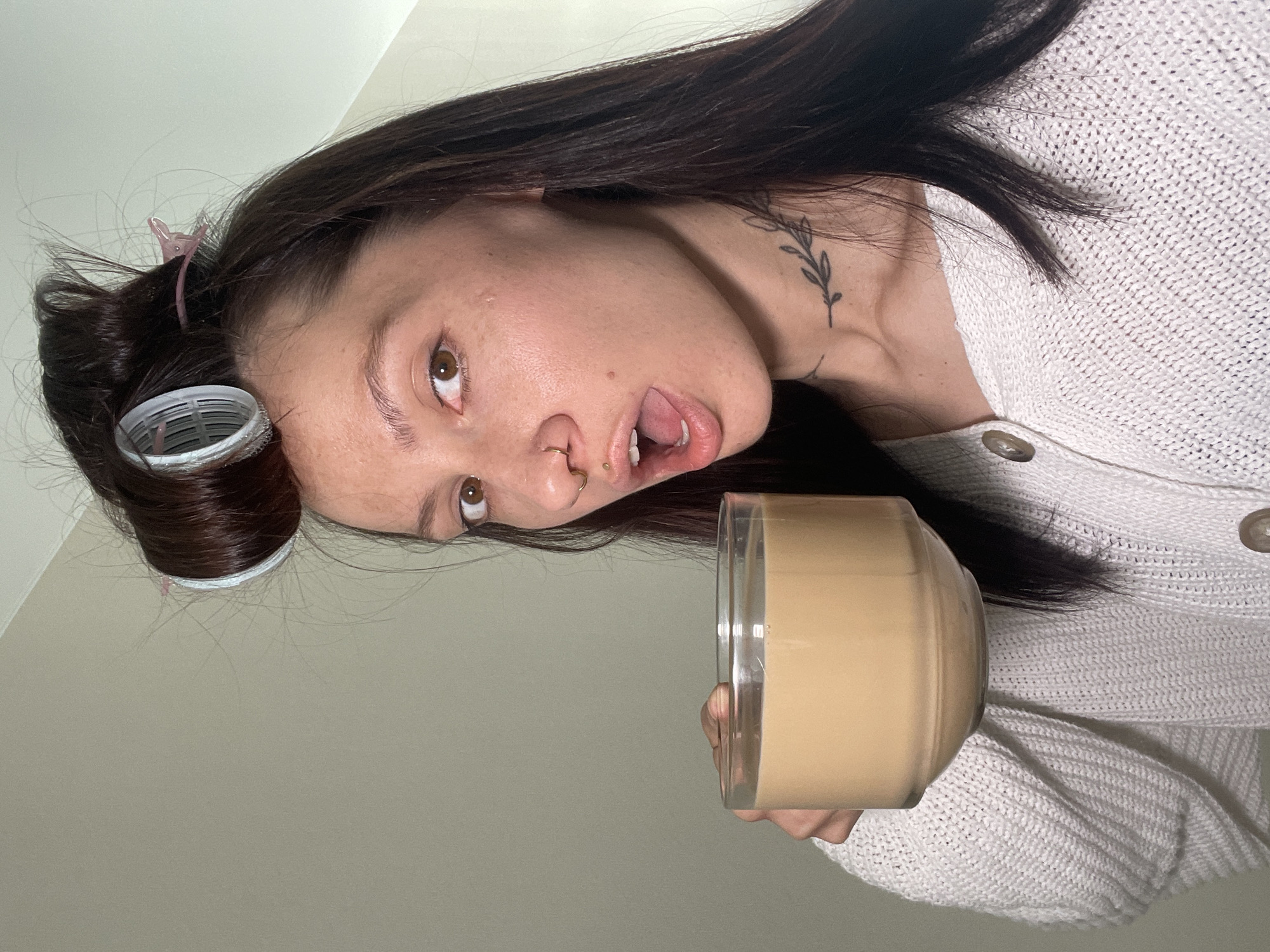 A casual indoor portrait features a young woman with long, dark brown hair, wearing a light-colored, possibly cream or white, knitted cardigan. Her head is tilted, and she looks directly at the camera with wide, dark brown eyes and her mouth slightly agape, suggesting a surprised or playful expression. Her hair is partially styled with a white hair roller visible on the left side of her head, secured with a pink hair clip.
Notable facial features include a small gold septum ring and a tiny gold labret piercing below her lower lip. A delicate black tattoo depicting a leafy branch is visible on the right side of her neck, just below her ear. She holds a clear glass or jar filled with a light brown, creamy beverage, likely coffee or a similar drink. Her hand is partially visible gripping the container.
The background consists of a plain, light grey or off-white wall, providing a minimalist setting for the portrait. The overall lighting is soft and even, typical of an indoor environment. The image itself is oriented vertically, with the woman's head appearing to be on its side. No specific location cues are visible within the photograph, but the provided context for the image is Izhevsk, Russia. There is no visible text.