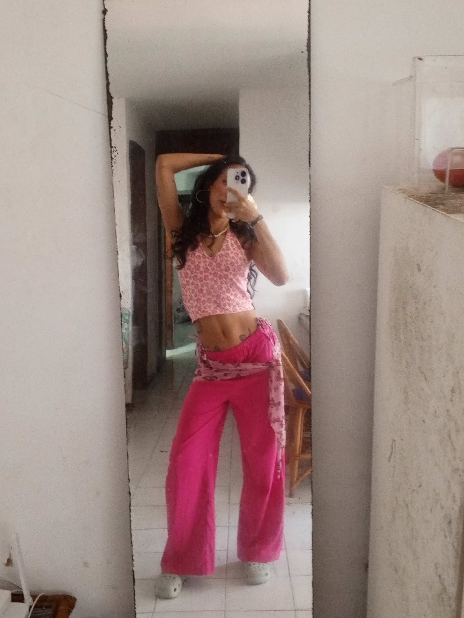 A young woman is standing in front of a full-length mirror in an indoor setting, likely a room in a home. She is holding her phone up, taking a selfie. The woman has dark, curly hair and is wearing a pink halter top with a floral pattern and bright pink wide-leg pants with a scarf tied around her hips. Her footwear appears to be Crocs. The room features white walls and a tiled floor. Behind her in the mirror's reflection are a doorway, a glimpse of another room, and a wooden chair. To the right of the mirror is a white cabinet-like structure with visible stains. The mirror itself has a dark, weathered frame. Based on the general architectural style and the possible subtropical appearance, the location might be Porlamar, Venezuela.