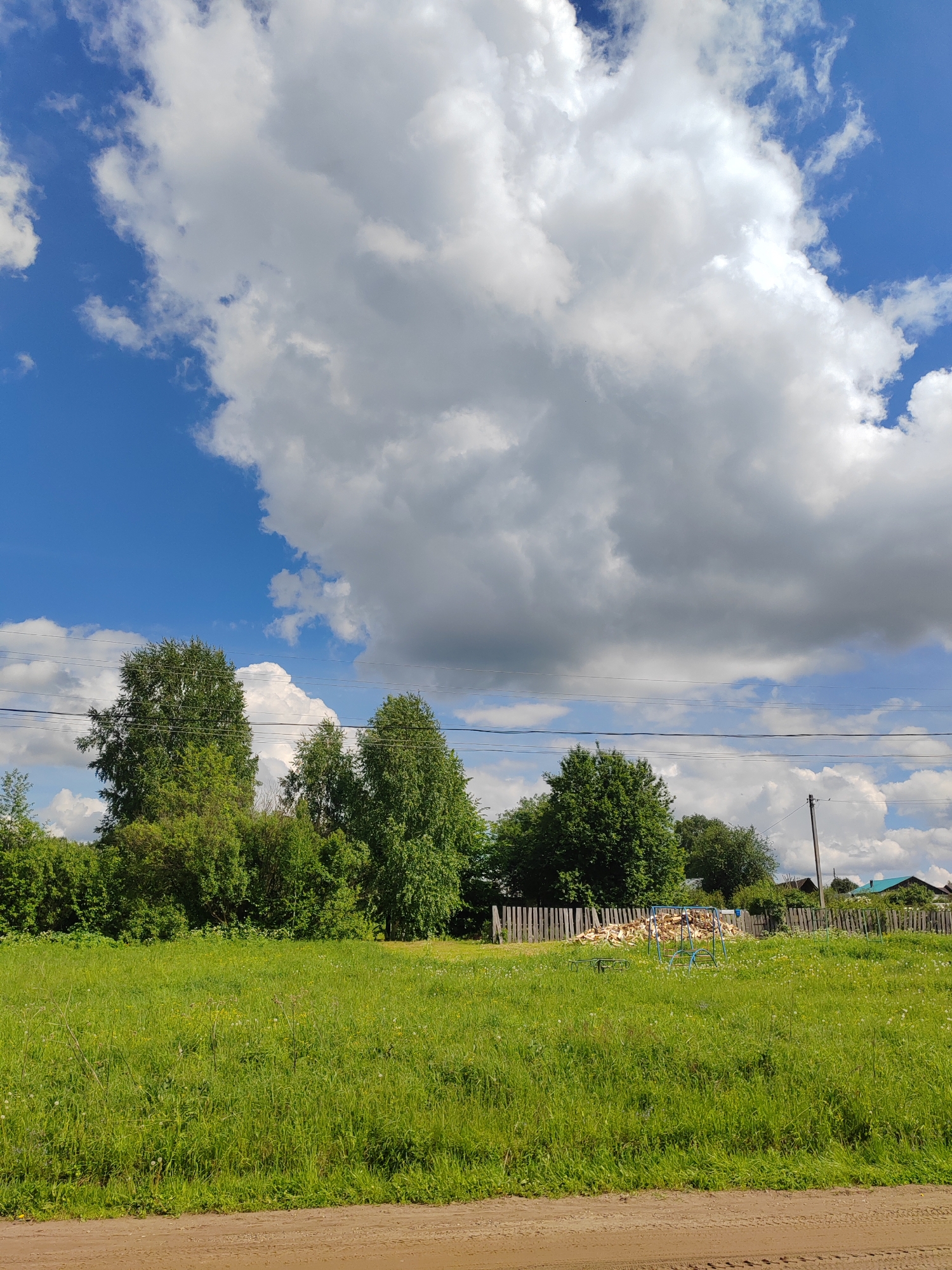 The image depicts a rural scene in Уральское, Russia. A large green field of tall grass stretches towards a line of trees and bushes in the midground. Behind the trees, a wooden fence is visible, with a pile of wood and some metal playground equipment situated in front of it. Power lines run horizontally across the scene above the trees. In the background, rooftops of houses with green roofs can be seen. The sky occupies the upper portion of the frame, featuring a vibrant blue sky partially obscured by large, puffy white clouds. The foreground consists of a dirt road.