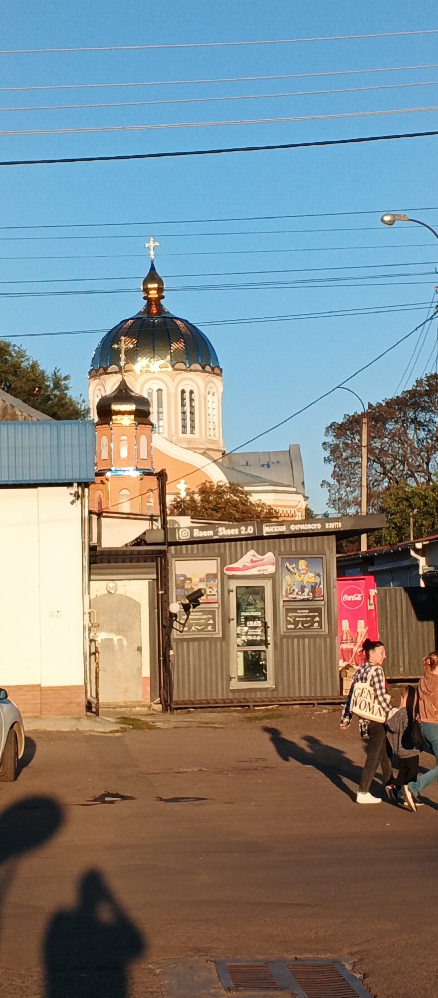 The image captures a street scene in Smila, Ukraine, with a church dome prominently featured against a clear blue sky. The church, adorned with a golden cross, appears to be an Eastern Orthodox church, indicated by its architectural style and onion domes. In the foreground, a small commercial structure with metal siding houses "Room Shoes 2.0," a shoe store, as evidenced by a Nike shoe advertisement on the facade and merchandise visible through the glass door. Coca-Cola branding is also visible on a cooler. Several individuals are walking on the asphalt road, casting long shadows due to the low angle of the sun, suggesting it is late afternoon or early morning. Power lines crisscross the sky. The general environment suggests an urban setting with a mix of residential and commercial buildings, and a prominent religious structure.
