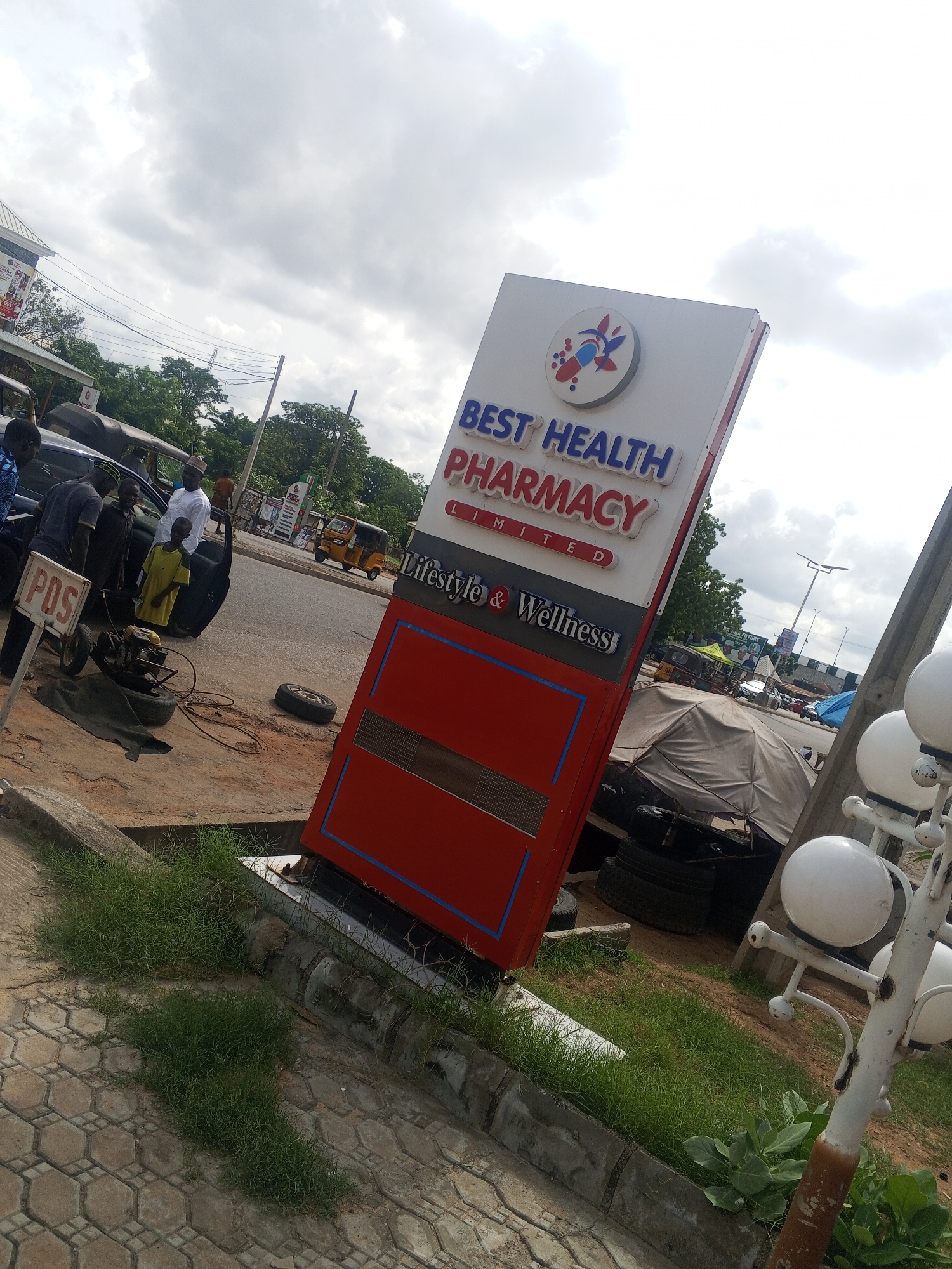 The image depicts a street scene in Jalingo, Nigeria, with a prominent red and white sign for "BEST HEALTH PHARMACY LIMITED" at the forefront, tilted slightly. Below the pharmacy name, "Lifestyle & Wellness" is visible. To the left of the sign, a group of men and a young boy are gathered around a dark-colored car, where a tire appears to be undergoing some work, possibly a repair or change, with a small engine or generator present on the ground. A "POS" sign is also visible on a pole near the car. In the background, a yellow tricycle, commonly known as an "okada" or "kekè," is driving on the road, alongside other vehicles and structures, including trees and buildings, under a cloudy sky. The general environment suggests a bustling urban setting.