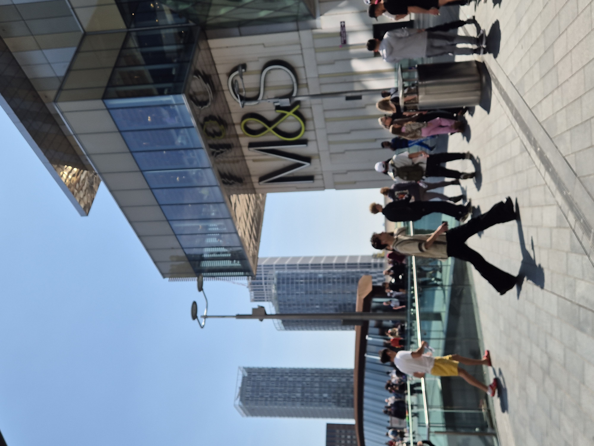 The image depicts an outdoor scene in London, United Kingdom, featuring a modern shopping area with numerous people. A prominent "M&S" (Marks and Spencer) store facade is visible, with people walking along the pavement in front of it. Several high-rise buildings are apparent in the background. A lamppost is positioned on the left side of the scene. The weather is clear and sunny.