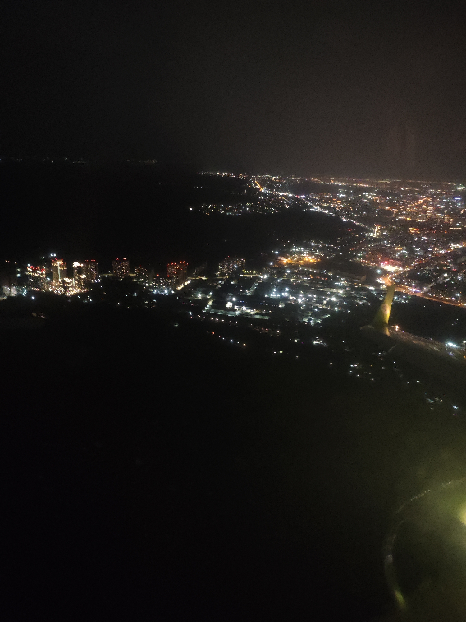 The image depicts an aerial view of a city at night, with city lights illuminating the landscape. The location is Novosibirsk, Russia. The foreground is dark, possibly representing an unlit area or a body of water. In the middle ground, there's a cluster of brightly lit skyscrapers. Further in the distance, the city lights stretch across the horizon, indicating the extent of the urban area. An airplane wing is visible in the lower right quadrant, suggesting the photograph was taken from an airplane.