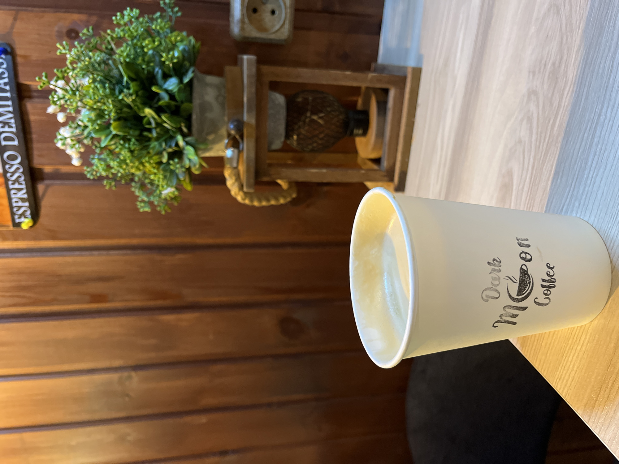 The image shows a coffee cup with the words "Dark Moon Coffee" on it. The cup is placed on a table with a light wood grain finish. In the background is a wall covered in horizontal wood panels. There's a small decorative plant and a wooden decorative object on the wall. Additionally, a sign that reads "Espresso Demitasse" vertically is visible on the wall. The scene's location in Petropavlovsk-Kamchatsky, Russia, cannot be confirmed from the image alone, as there are no distinctive landmarks or cultural elements indicating that location.