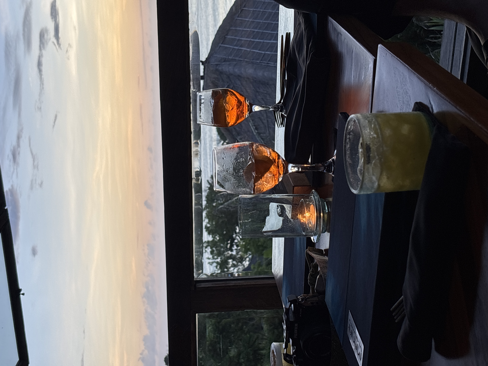 The image shows a view from a restaurant in Kuta Selatan, Indonesia at sunset. Through a window, the sky is visible with orange and yellow hues. On the table inside are two glasses of Aperol Spritz, a glass of a yellow beverage (possibly a cocktail), a lit candle in a glass holder, silverware wrapped in dark napkins, a Fujifilm camera, and a table with a dark blue surface. The edge of the table is wood.