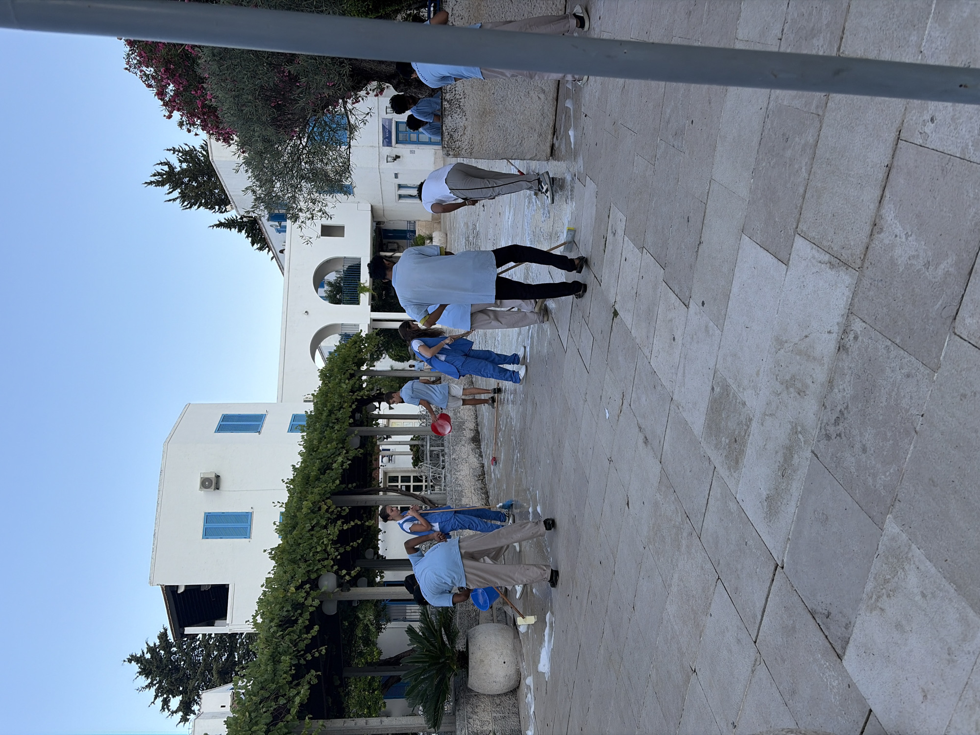 In Budva, Montenegro, several individuals dressed in light blue uniforms are actively engaged in cleaning a paved outdoor area. The scene appears to be part of a resort or hotel complex, characterized by white buildings with blue shutters and lush greenery. The individuals are using mops and buckets, with visible suds indicating they are washing the stone-tiled ground. Some are bending over their cleaning tasks, while others stand or move across the area. The environment is bright with natural light, suggesting it is daytime. The overall impression is one of collective effort to maintain the cleanliness of the public space within this Mediterranean setting.