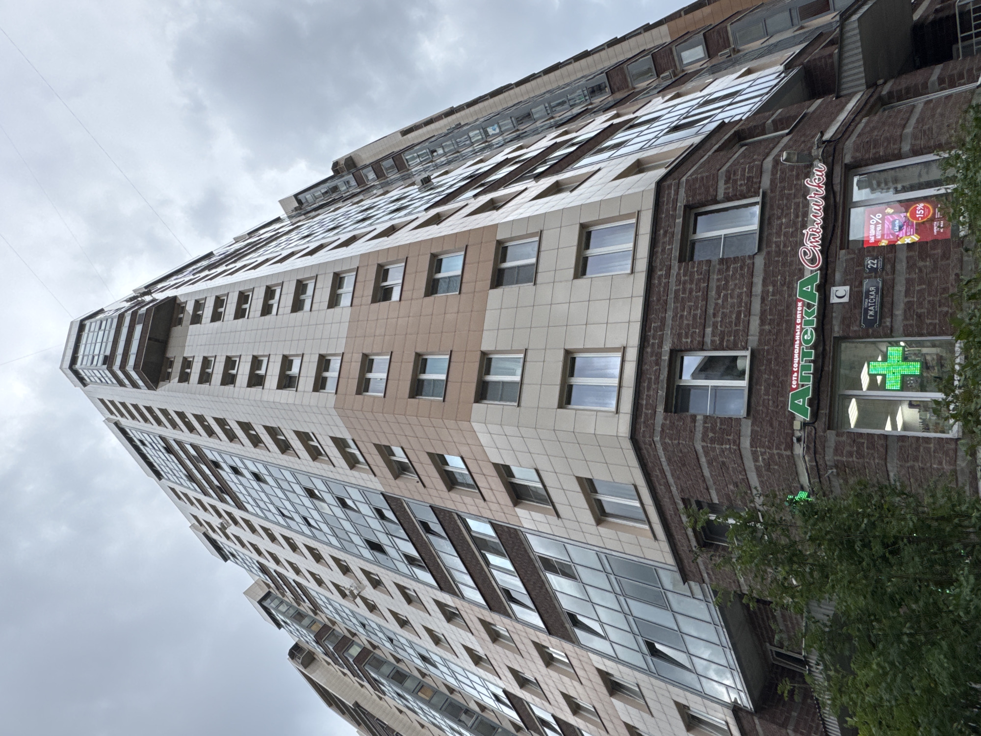 The image captures a low-angle view of a multi-story modern apartment building against an overcast sky. The building features a mix of light-colored tiled facades and extensive glass windows, with balconies on some levels. The lower section of the building is clad in dark brick. At street level, a pharmacy with illuminated green cross signage and a neon sign reading "Аптека Столичка" is visible, along with various advertisements in store windows. The pharmacy is located at Gzhatskaya Street, building number 22, in Sinyavino, Russia. Green foliage from trees partially obscures the lower part of the building in the foreground. There are no people visible in the frame.