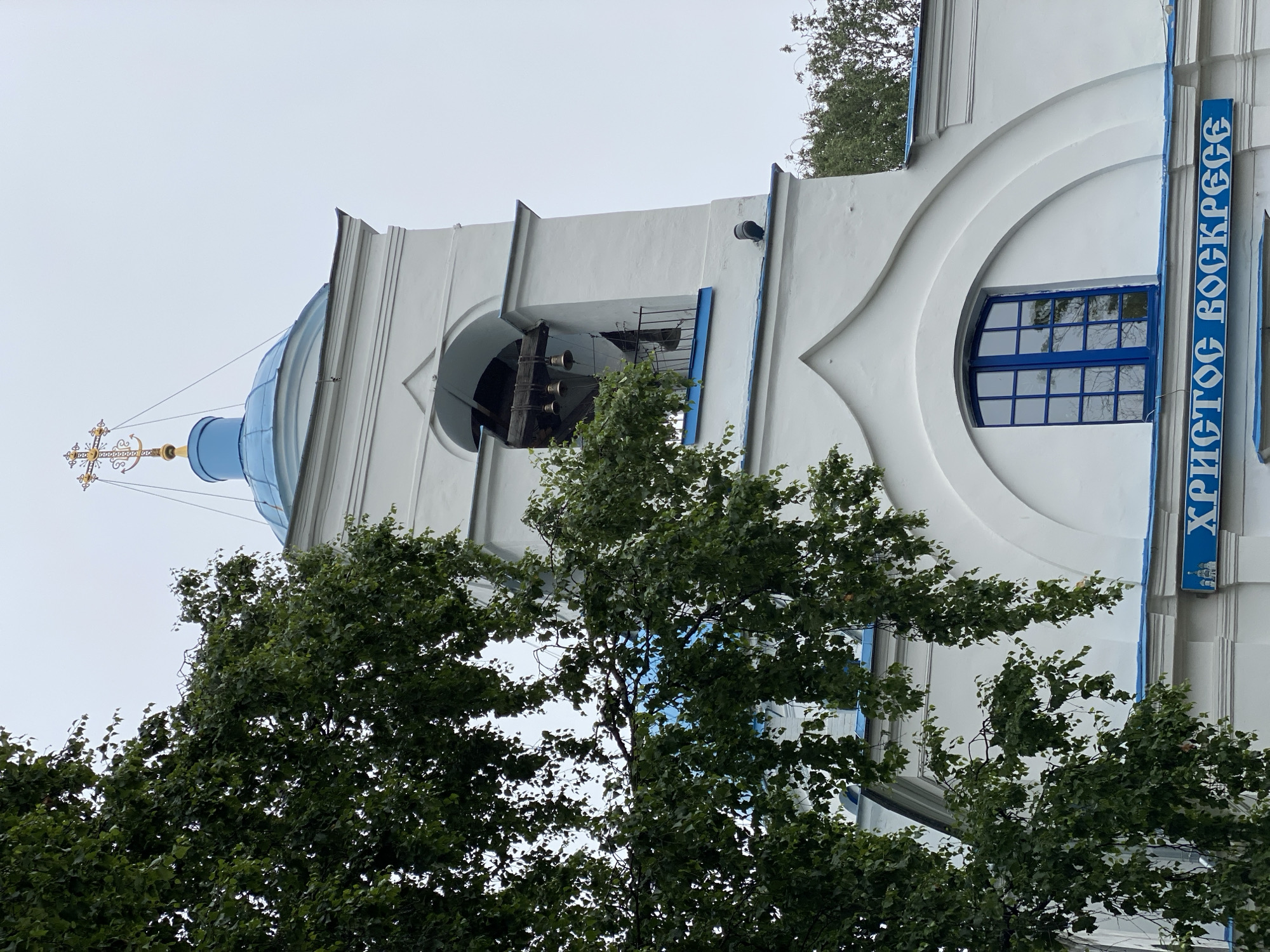 The image shows the facade of a white church with blue accents. A prominent feature is a bell tower with a blue dome, topped by a golden cross. The tower has arched openings where several bells are visible, along with a small railing. Below the bell tower, a arched window with blue panes is set into the white wall, and a blue sign with white Cyrillic lettering that reads "ХРИСТОС ВОСКРЕСЕ" is mounted below the window. Lush green foliage from a tree partially obscures the left side of the church. The sky is overcast. The church is located in the city of Priozersk, Russia.