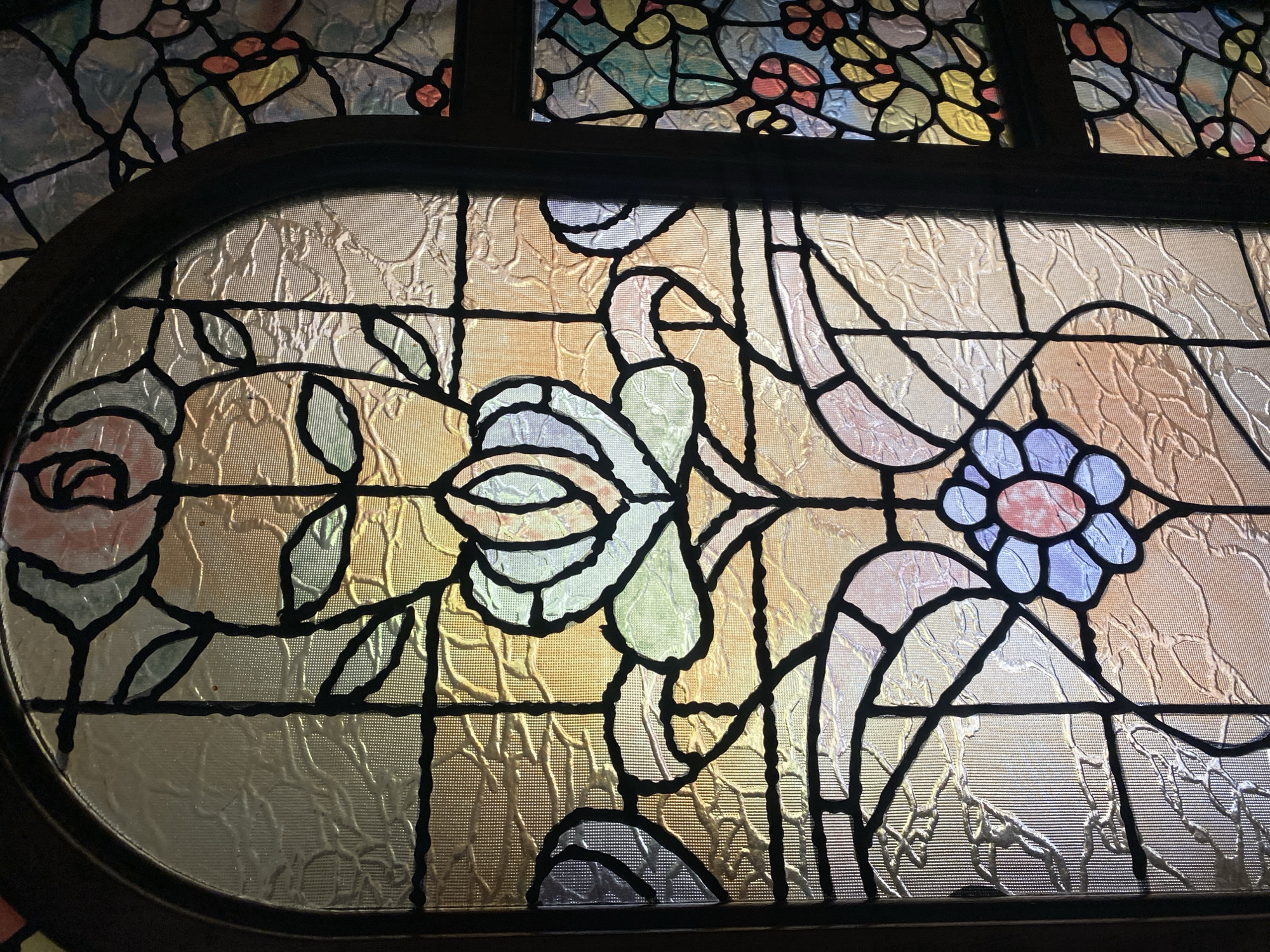 The image shows a close-up of a stained-glass window, likely interior, with floral motifs in an oval frame. The glass has a textured, slightly frosted appearance. The flowers are outlined in black lead or a similar material. A rose, colored in shades of pink, white, and green, is situated on the left side. In the middle of the window is a light-colored flower. A blue flower with a pink center is on the right. The background glass is predominantly a warm golden color. Above the oval section is a row of colored glass panels. The location is identified as Yekaterinburg, Russia.