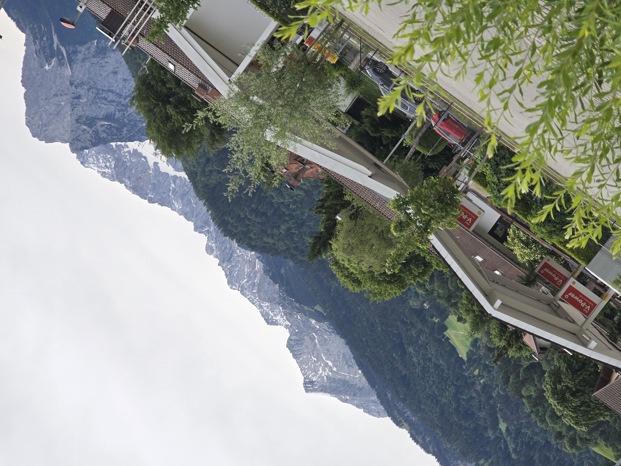 The image captures a picturesque view of a town nestled within a majestic mountainous landscape, indicative of Garmisch-Partenkirchen, Germany. In the background, towering mountains stretch across the horizon, their higher elevations dotted with patches of snow. The lower slopes are densely covered in dark green coniferous forests, which extend into the mid-ground. A small patch of bright green pasture is visible on a hillside in the forested valley.
The foreground features several buildings with traditional reddish-brown tiled roofs and white facades, some equipped with balconies. On the right side, signs for a Shell service station are clearly visible, displaying "Shell" and "V-Power Performance Fuel" in red and white. A paved road runs in front of these buildings, where a red car and a dark-colored car are parked. Lush green foliage, including the leaves of a foreground tree, partially frames the scene. The sky is uniformly overcast and light grey, suggesting a cloudy daytime. No specific activities or people are visible, contributing to the tranquil alpine atmosphere of the scene.