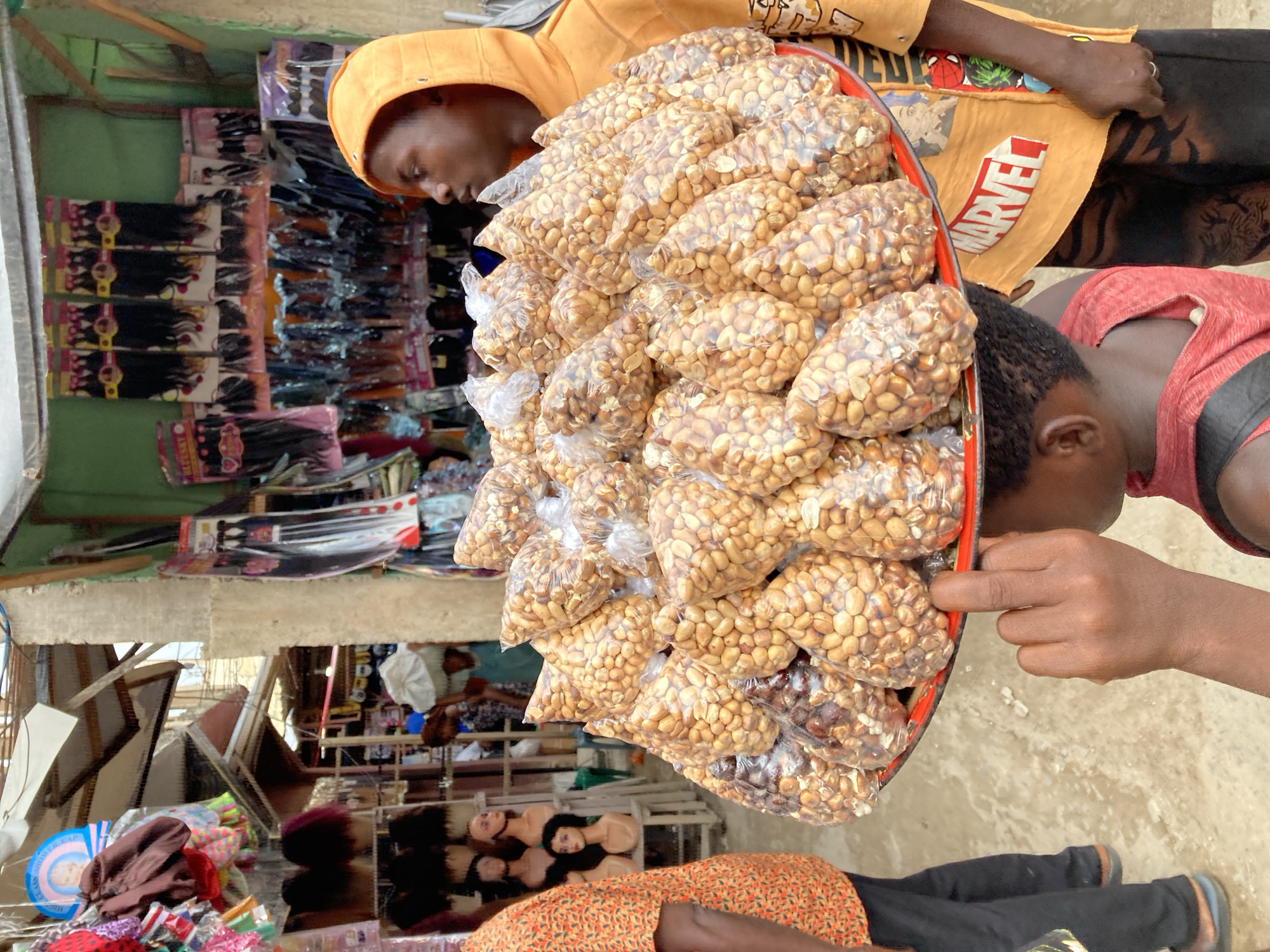 The image shows an outdoor scene in Ura Nta Umuarandu, Nigeria. Two individuals are visible, one wearing an orange Marvel-themed shirt and the other a red shirt. They are standing or sitting near a display of packaged peanuts in clear plastic bags, arranged on a red tray. The peanut display is the central focus. In the background, there are various commercial items including packaged hair extensions and wig heads. The setting appears to be a market or street-side vending location.