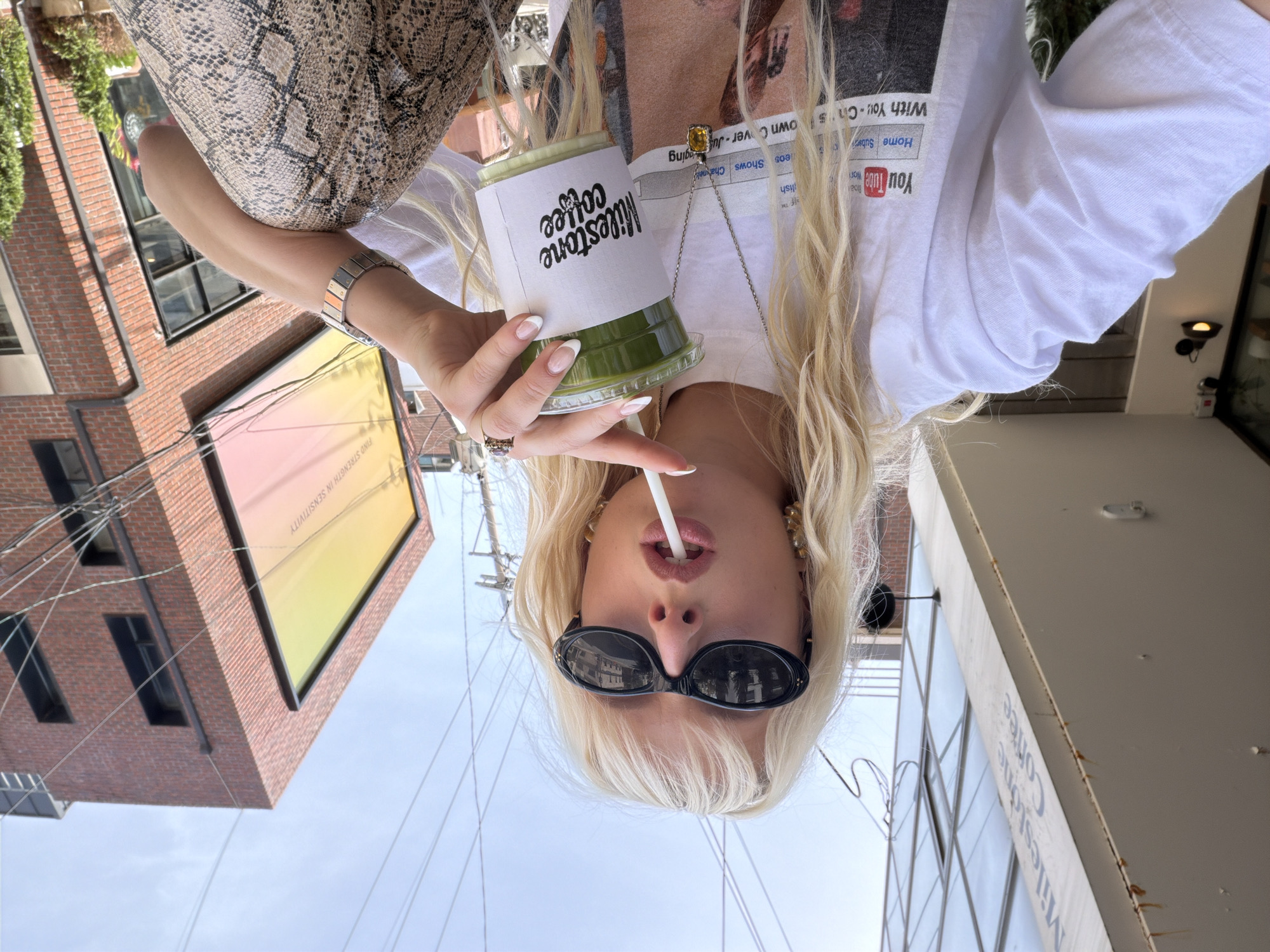 A blonde-haired woman wearing sunglasses sips a drink with a straw outdoors. The drink is in a cup labeled "Milestone Coffee" with a white sleeve. The woman is wearing a white t-shirt with a YouTube logo and a necklace. She has an animal print purse on her shoulder. The background shows a brick building and utility lines. The drink is green and appears to be a green tea latte. The location is identified on the sign as being in the city of Seoul, South Korea.