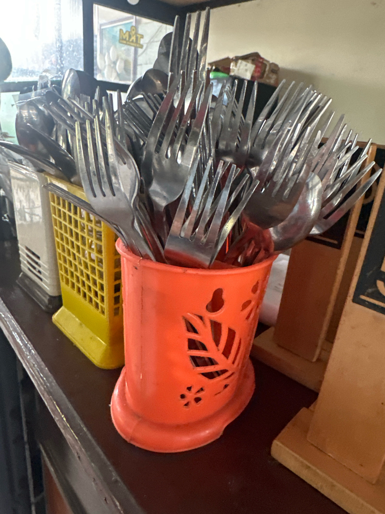 The image shows a close-up of an orange plastic utensil holder filled with various metal cutlery, predominantly forks and some spoons. The holder has a decorative cut-out pattern of leaves and flowers. To the left of the orange holder is a yellow plastic basket-like container, also holding cutlery. Further to the left, partially visible, is a white rectangular object with horizontal slits. The background includes a blurry window with a sign that appears to say "MAT", and behind it, some boxes are stacked. The overall scene suggests a food service environment, likely a restaurant or a warung, possibly in Mojokerto, Indonesia, given the provided context. The lighting is somewhat dim, with reflections visible on the metal cutlery.