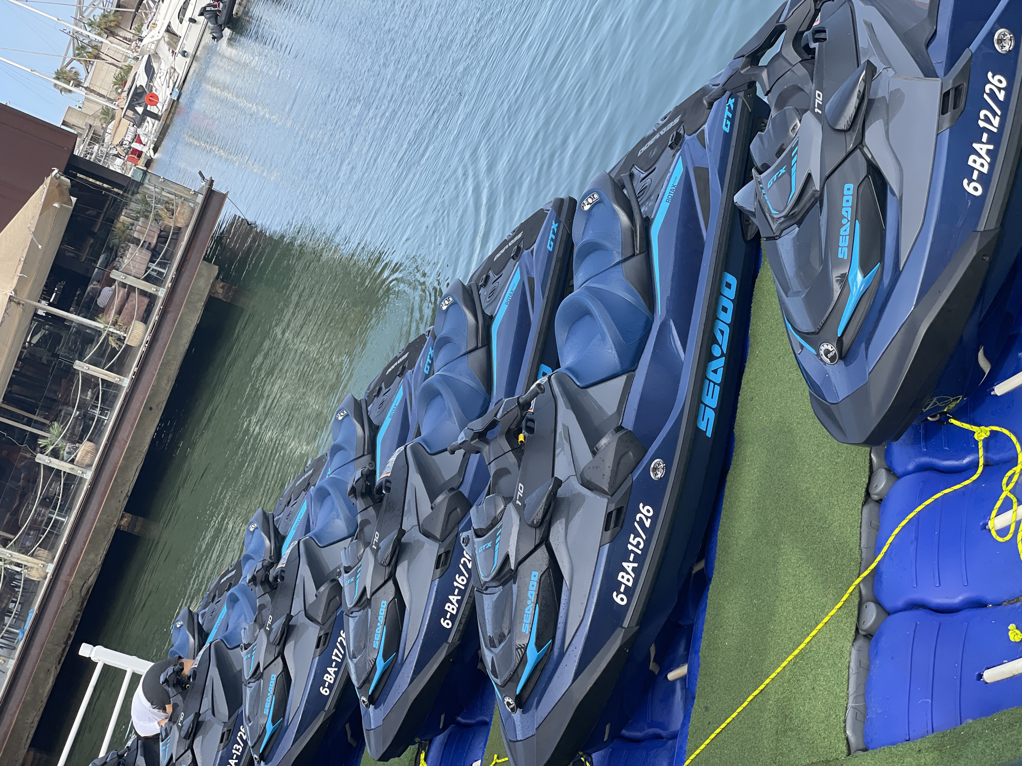 This image depicts a row of navy blue Sea-Doo jet skis docked on a waterway. The jet skis are lined up next to a floating dock covered in green astroturf, with blue plastic pontoons visible underneath. A person wearing a white shirt and a dark hat is partially visible, standing on the dock near the jet skis.
Several jet skis have visible text, including "ROTAX", "GTX", and "SEA-DOO", along with alphanumeric identifiers such as "6-BA-17/25", "6-BA-16/2", "6-BA-15/26", and "6-BA-12/26". The background shows a body of water with ripples, suggesting a calm day. In the upper left corner, there are glimpses of boats and buildings, hinting at a marina or waterfront area. The lighting suggests it is daytime. The overall scene is one of organized watercraft ready for use or rental in a sunny, possibly coastal, environment.