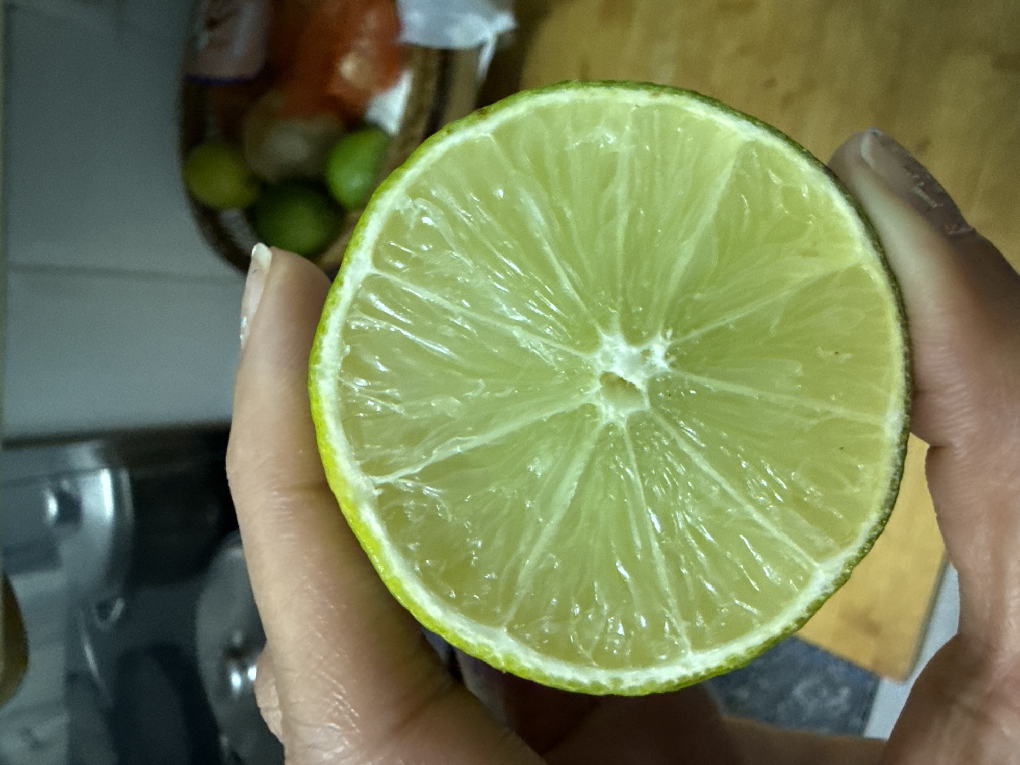 A close-up shot features a hand holding a freshly cut lime against a softly blurred background, likely an indoor kitchen setting. The main subject is half a lime, revealing its vibrant green rind and the translucent, pale green, juicy pulp divided into distinct segments radiating from the center. The hand, with visible skin texture, firmly holds the lime, with fingers and thumb partially visible.
In the out-of-focus background, a container or basket is visible, holding several small green objects, likely more limes or similar produce, along with some indistinct red or orange items. Parts of a kitchen counter and metallic kitchen fixtures, possibly a sink or faucet, are also faintly discernible. The lighting suggests an indoor environment, and no specific time of day or weather can be determined. There is no visible text in the image. The scene captures the lime in detail, suggesting readiness for culinary use or drink preparation.