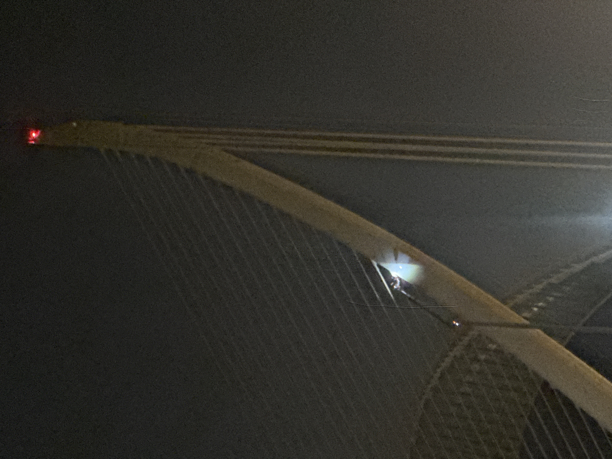 This is a low-light, slightly grainy, nighttime photograph of a portion of the Puente de las Flores, a cable-stayed bridge located in Valencia, Spain. The bridge's main pylon, a tall, cream-colored structure, dominates the right side of the frame, tapering towards its apex where a red navigation light is visible. Numerous thin, pale cables, which are part of the bridge's structure, extend diagonally across the image, creating a striped effect against the dark sky.
In the middle-left portion of the image, a single individual is visible. They are positioned on a section of the bridge's walkway or superstructure, leaning forward and appearing to be working or conducting an inspection. A bright, focused light source, likely a headlamp or spotlight, is directed downwards from the individual, illuminating a small area of the bridge. This light creates a distinct bright spot and a subtle halo effect. Below and to the left of this person, a small orange-red light is also visible, possibly another navigational marker or a worker's signal light. The rest of the scene is dark, with the surrounding environment obscured by the night and potentially fog or mist, which diffuses the lighting and contributes to the overall atmospheric, somewhat mysterious quality of the image.