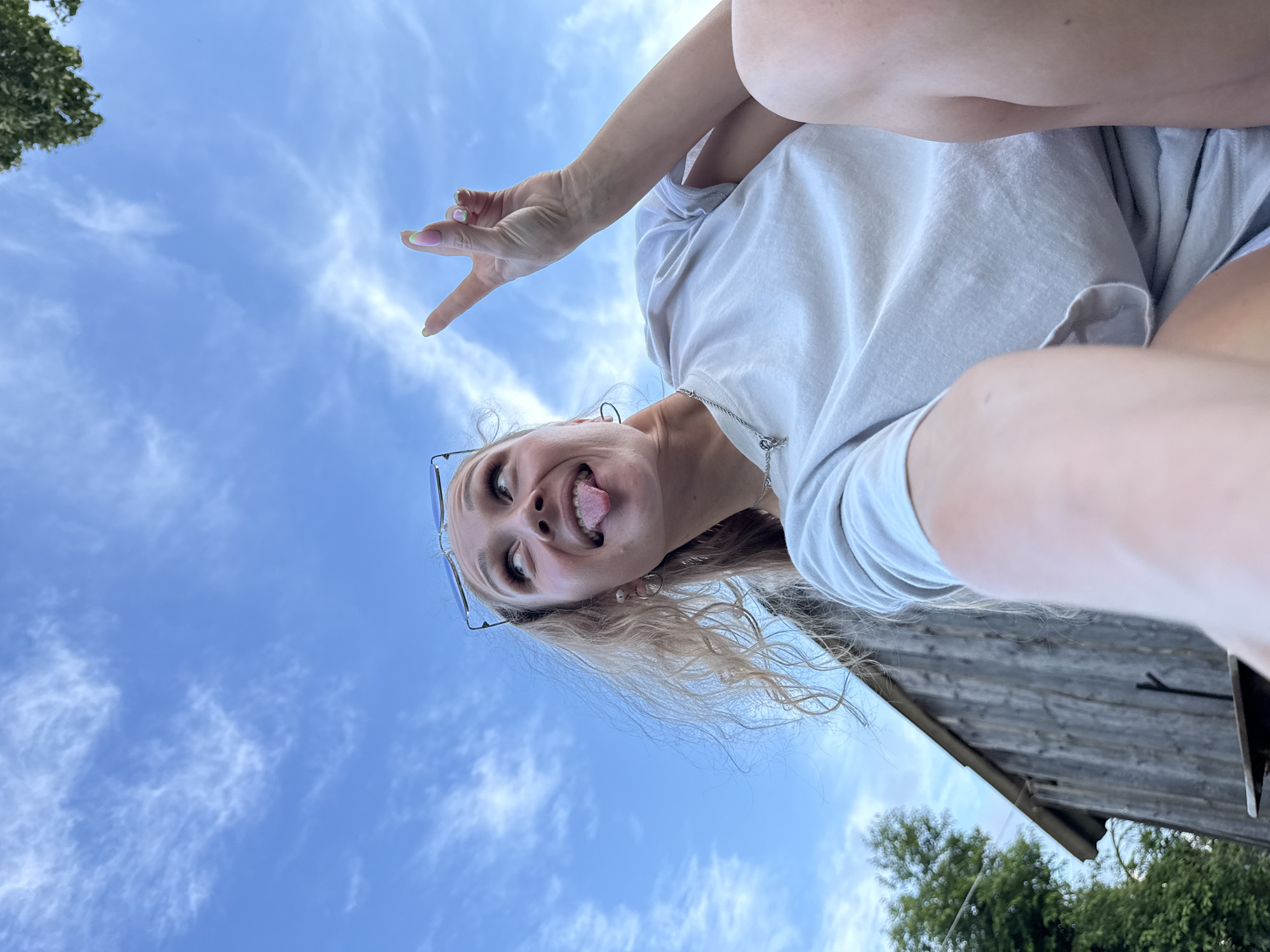 A young woman is captured in a low-angle, upside-down selfie against a bright blue sky with wispy white clouds. She has blonde hair and is wearing a light-colored t-shirt. Her left hand is raised, forming a peace sign. She is sticking her tongue out at the camera and appears to be in a playful or mischievous mood. Her sunglasses are perched on her head. In the background, parts of a rustic wooden structure and green foliage are visible, suggesting an outdoor setting. The lighting indicates it is daytime.