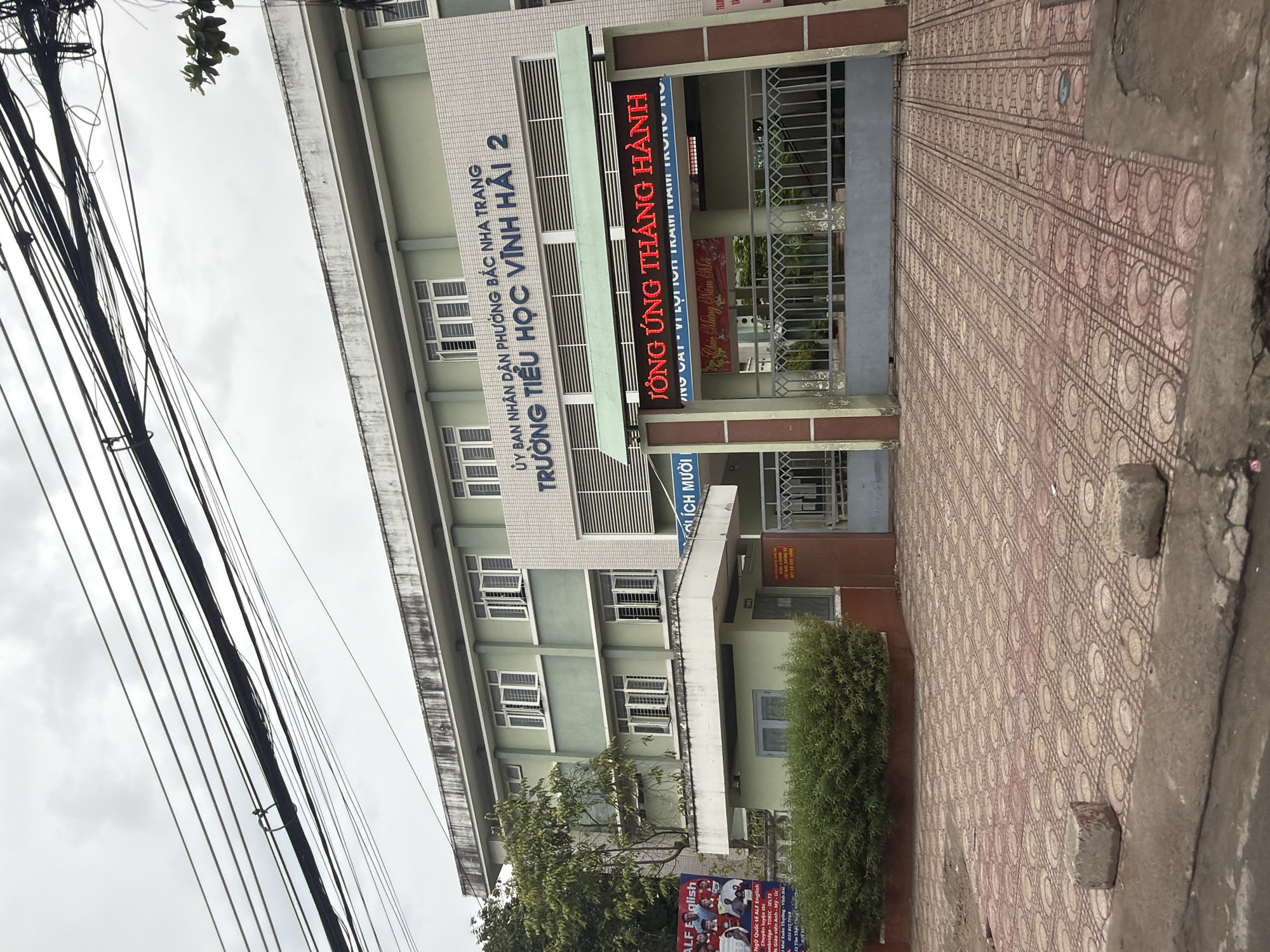 The image shows the front exterior of a light green, multi-story building, identified by signage as "TRƯỜNG TIỂU HỌC VĨNH HẢI 2" (Vinh Hai 2 Primary School). The building has a modern design with multiple rows of windows, some of which are open. Above the main entrance, a digital LED sign displays text in red, reading "TỔNG ỨNG THÁNG HÀNH" (General Application Month). To the left of the entrance, a banner with images of children and text in Vietnamese, including "ALF English," is visible. The building is situated on a street with a patterned tiled sidewalk and a concrete curb in the foreground. Overhead, a complex network of electrical wires crisscrosses the sky, partially obscuring the building. Lush green bushes line the base of the building in front of the sidewalk. The overall scene suggests an educational institution in an urban environment, possibly during a daytime with overcast skies.