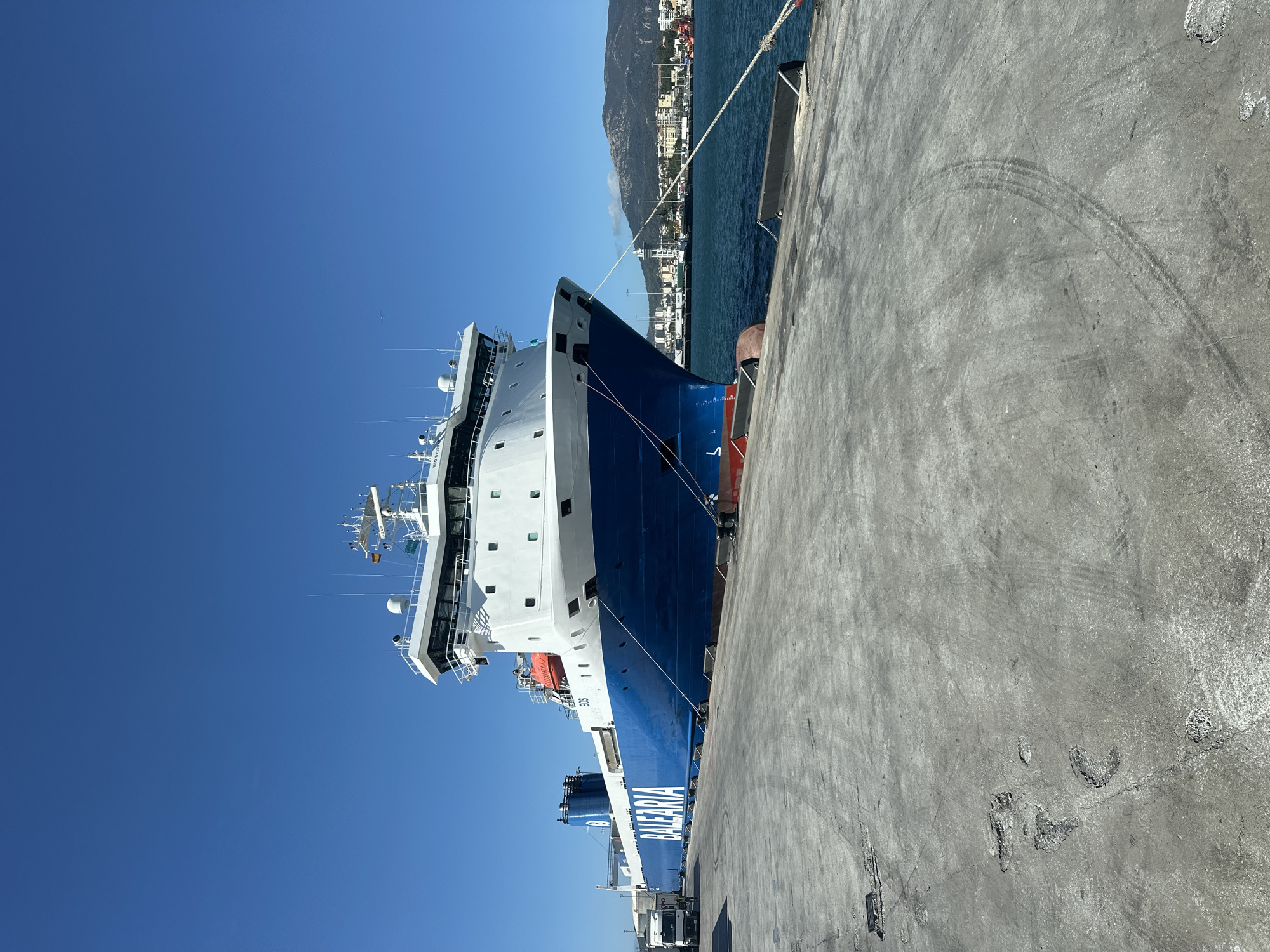 A large blue and white ferry named "BALEARIA" is docked at the port of Algeciras, Spain. The ferry is prominently featured in the foreground, taking up a significant portion of the image. It is tied to the dock with thick ropes. To the left of the ferry, a white truck is visible, suggesting commercial activity. The dock itself is made of concrete and shows signs of wear, with tire marks and cracks. In the background, the city of Algeciras unfolds with buildings and infrastructure lining the waterfront, backed by a hazy mountain range under a clear blue sky. The sea is calm and a deep blue.