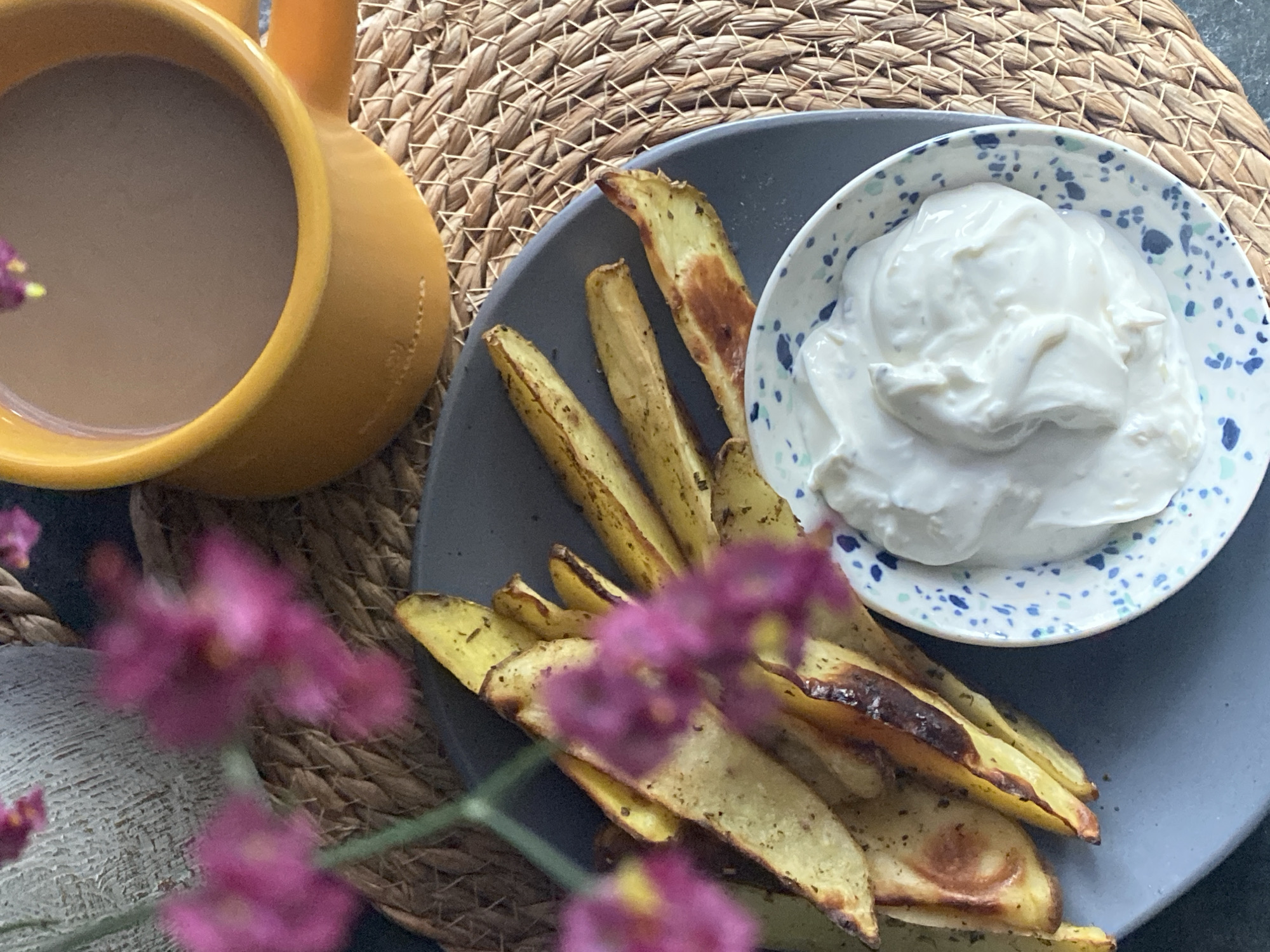 The image shows a plate of baked potato wedges with a small bowl of dip, a yellow mug of coffee, and pink flowers, all arranged on a woven placemat. The scene is likely indoors, with a neutral-colored wall in the background. The lighting is soft and natural. There is no indication of the city of Chaikovsky, Russia, or any specific geographical location.