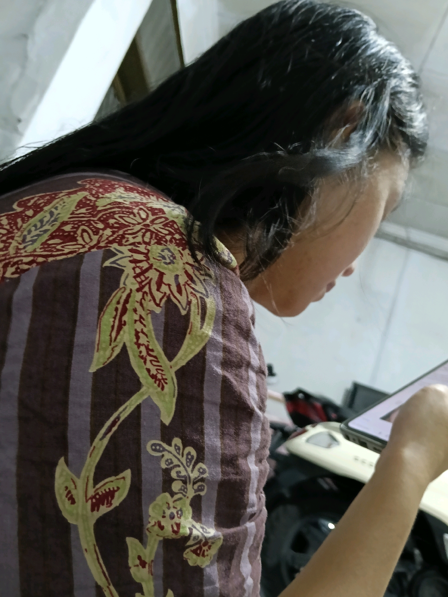 In the city of Jatimulya, Indonesia, a close-up, side-profile shot captures a person with long, dark hair engrossed in a mobile device. The individual is wearing a brown and purple striped shirt adorned with a traditional batik pattern featuring red and light green floral motifs. The person's right arm and hand are visible, holding and interacting with a smartphone, which displays a bright screen. The background is blurred, but suggests an indoor or covered outdoor setting with hints of white surfaces and dark objects, possibly vehicles or furniture. The lighting appears to be from overhead, casting a subtle glow on the person's face and hair.