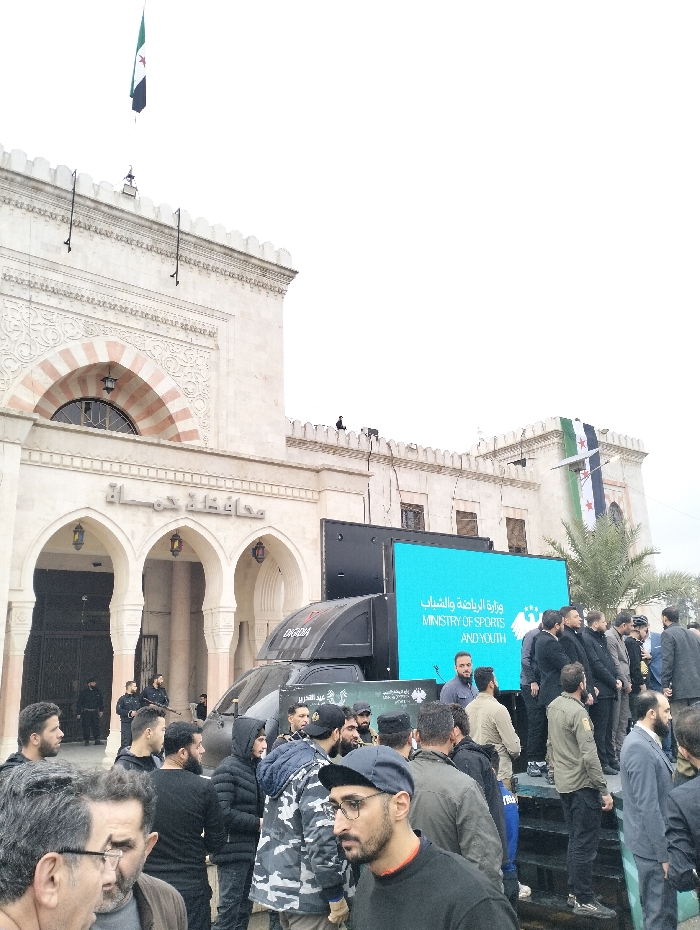 A crowd gathers outside a building with Syrian flags displayed. The building has arched windows and detailed stonework. A large truck with a digital screen displaying “Ministry of Sports and Youth” in Arabic and English is parked nearby. Men in various attire, including casual wear and uniforms, stand in front of the building. Security personnel with rifles are visible inside the building's entrance. The sky is overcast.
