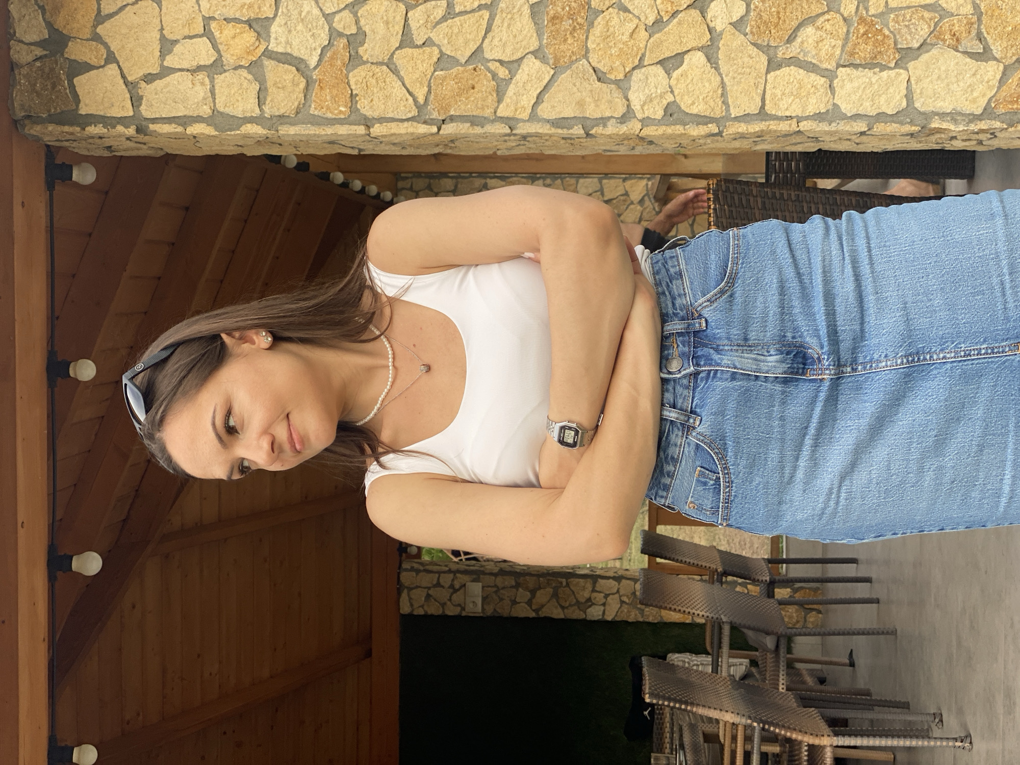A woman with brown hair and brown eyes, wearing a white tank top, a pearl necklace, a silver watch, and a denim skirt, stands with her arms crossed and looks to her left. She is standing on a paved area in front of a building with a stone exterior and a wooden roof. There are tables and chairs to her left. The location is Molovata Nouă, Moldova.