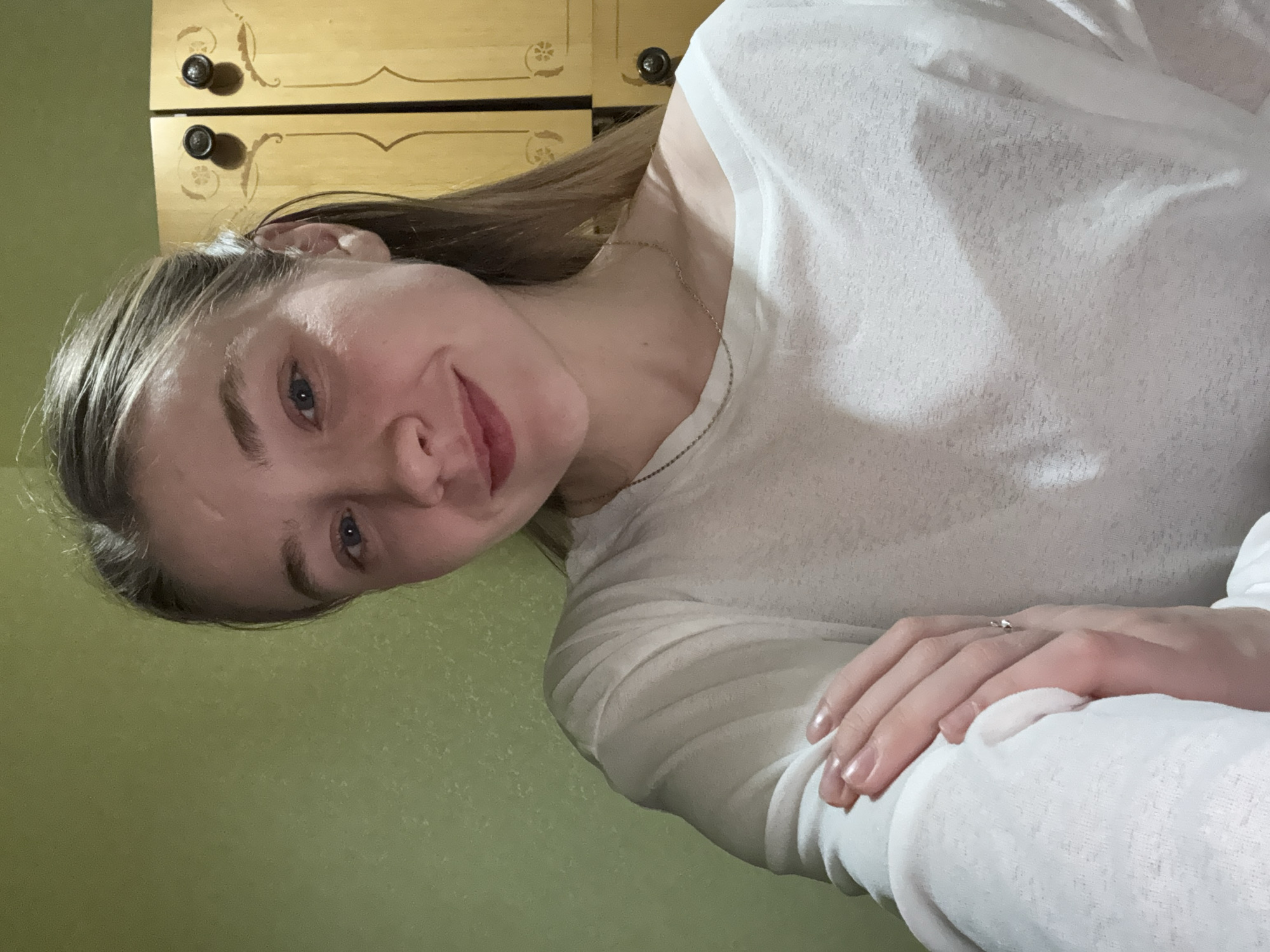 A fair-skinned young woman is photographed indoors, angled to the left. She is wearing a light-colored, long-sleeved shirt and a delicate necklace. Her hands are visible, folded over her arm, and a ring is on one finger. The wall behind her is a muted green. Behind her head is a wooden dresser with two visible drawers, each having a decorative pull. The location cannot be determined to be Izhevsk, Russia from the visual evidence.